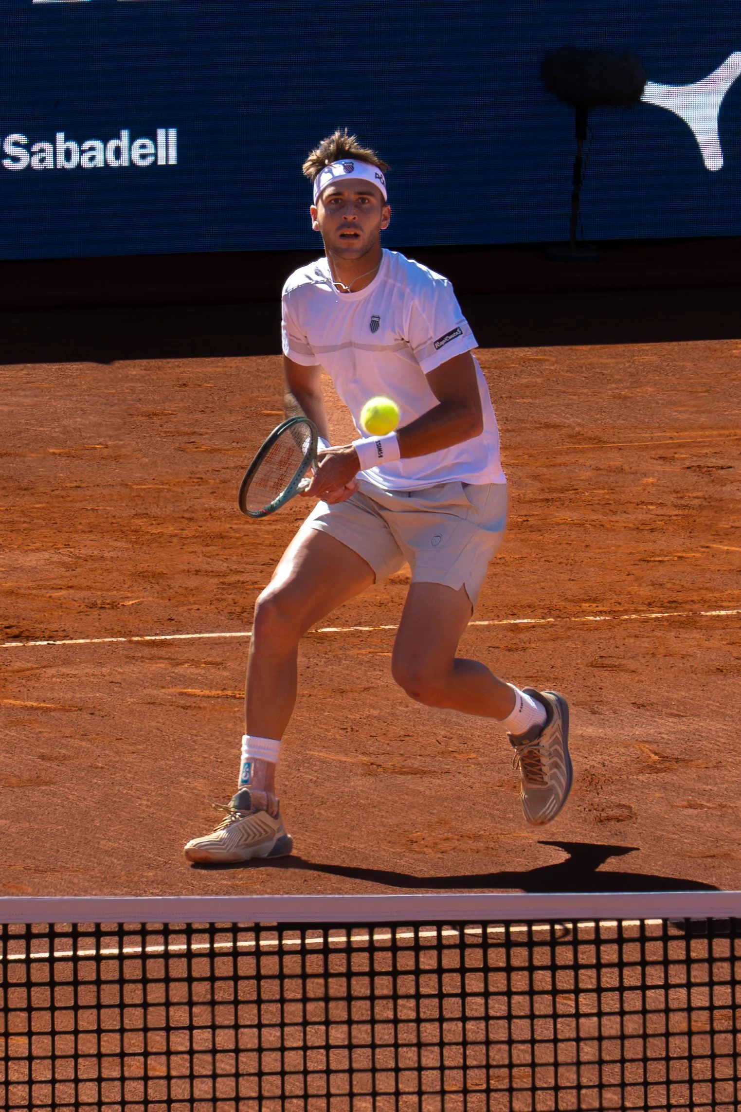 Tomas Martin Etcheverry on a clay court, wearing a white shirt, white shorts, a white headband, and sports shoes, preparing to hit a tennis ball.