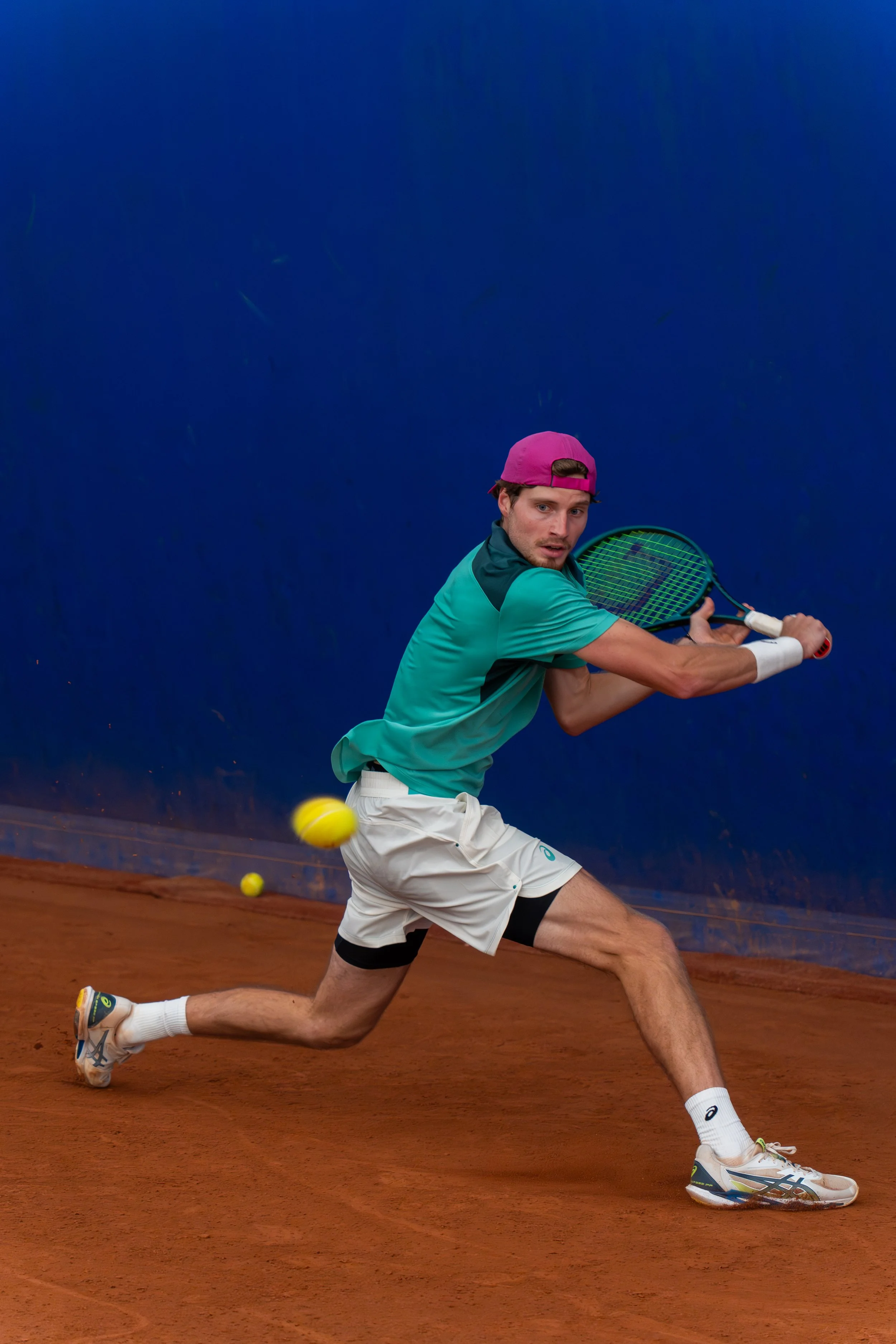 A male tennis player in teal shirt, white shorts, and a pink cap, is lunging to hit a tennis ball on a red clay court.