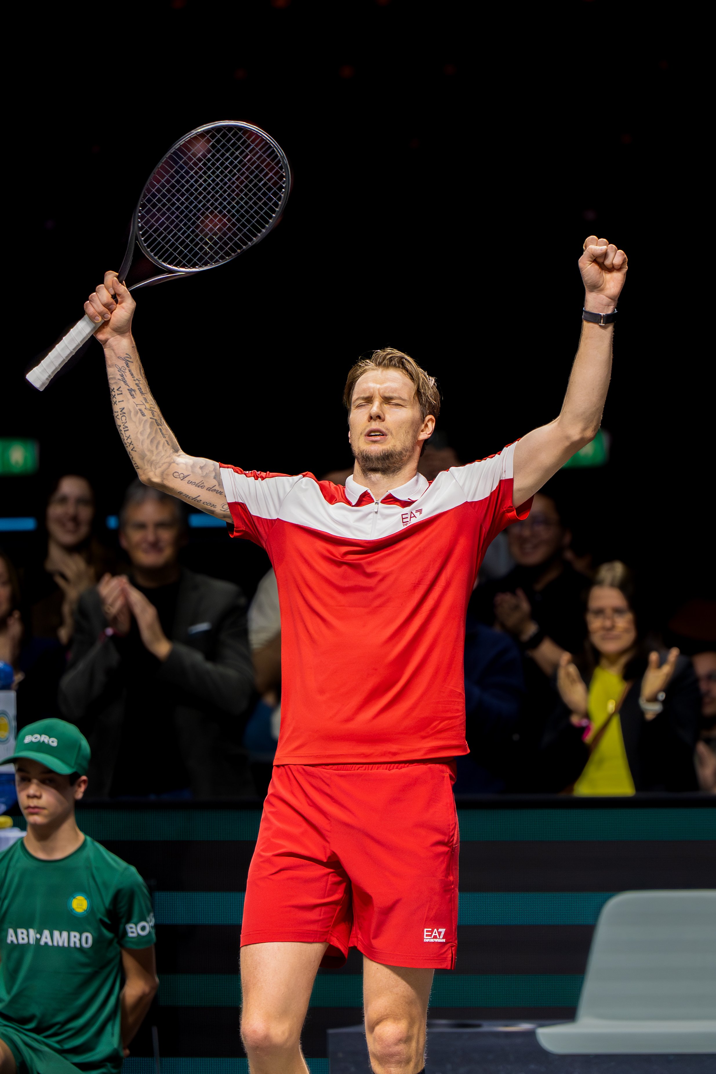 Alexander Bublik celebrates with his arms raised, holding a tennis racket in his right hand and his eyes closed. People in the background are clapping and smiling.