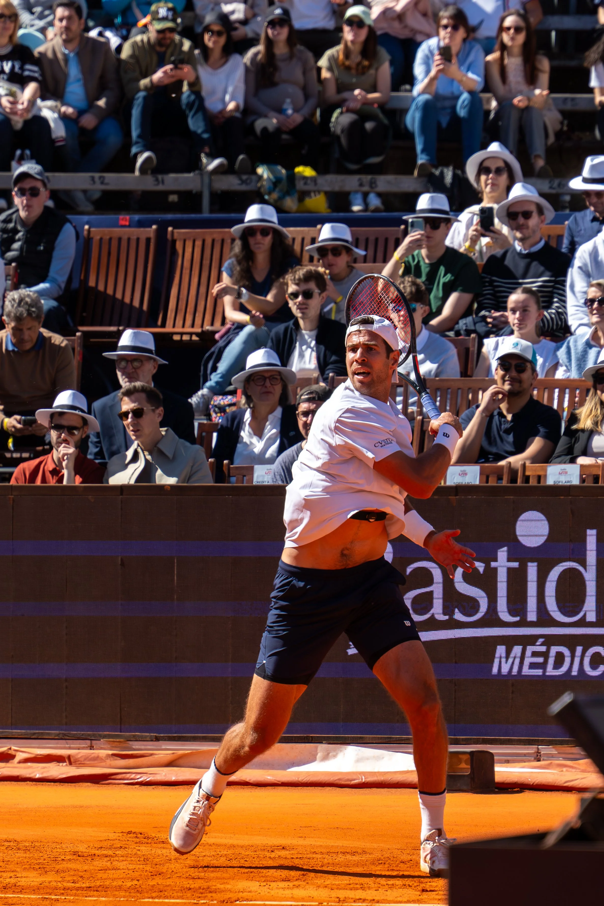 A male tennis player in a white shirt and black shorts playing on a clay court during a match, with a crowd of spectators in the background watching. Some spectators are eating, wearing sunglasses, and holding cameras or phones, with several wearing 