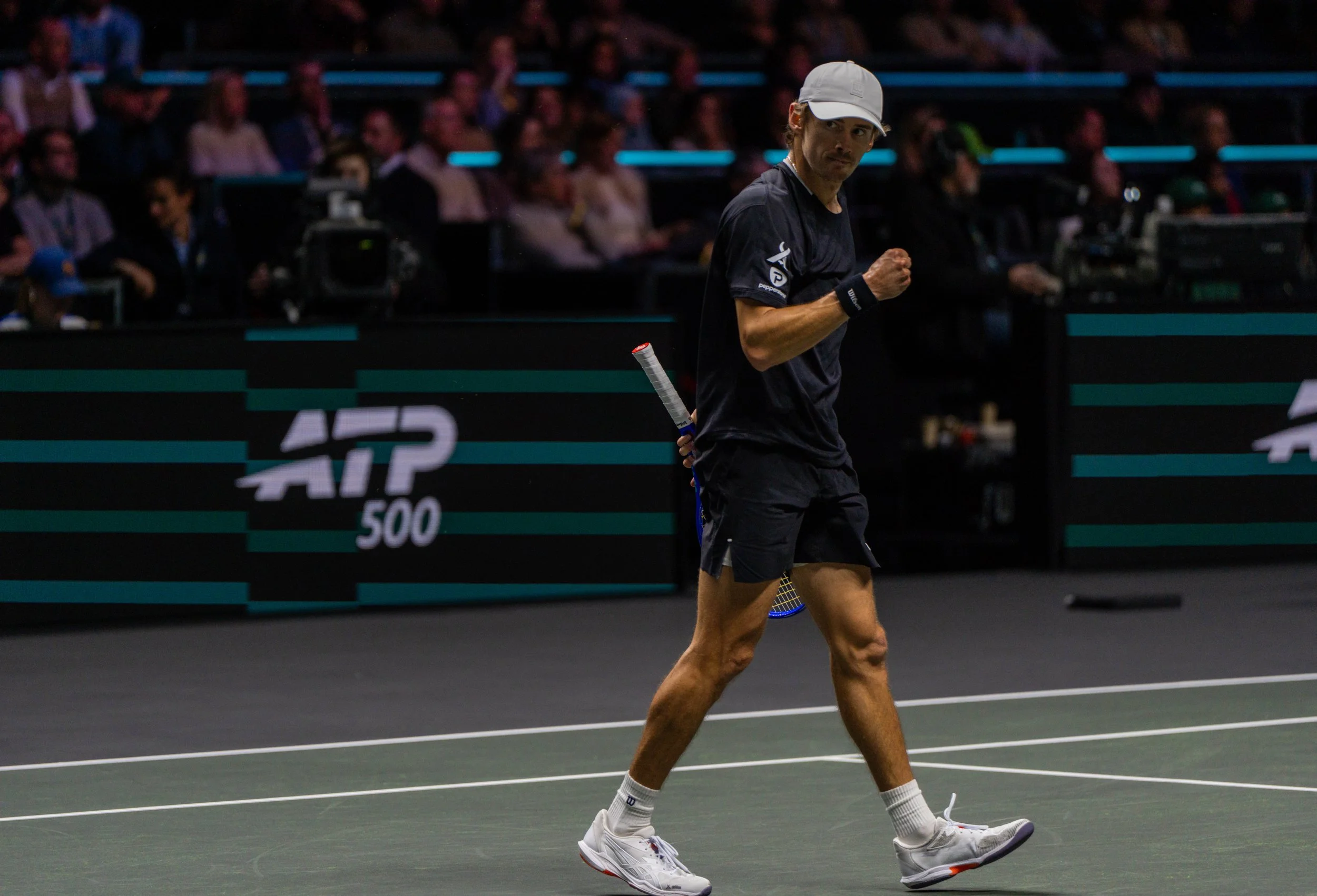 Alex de Minaur, holding a tennis racket, celebrating on a tennis court with a crowd in the background during an ATP 500 event.