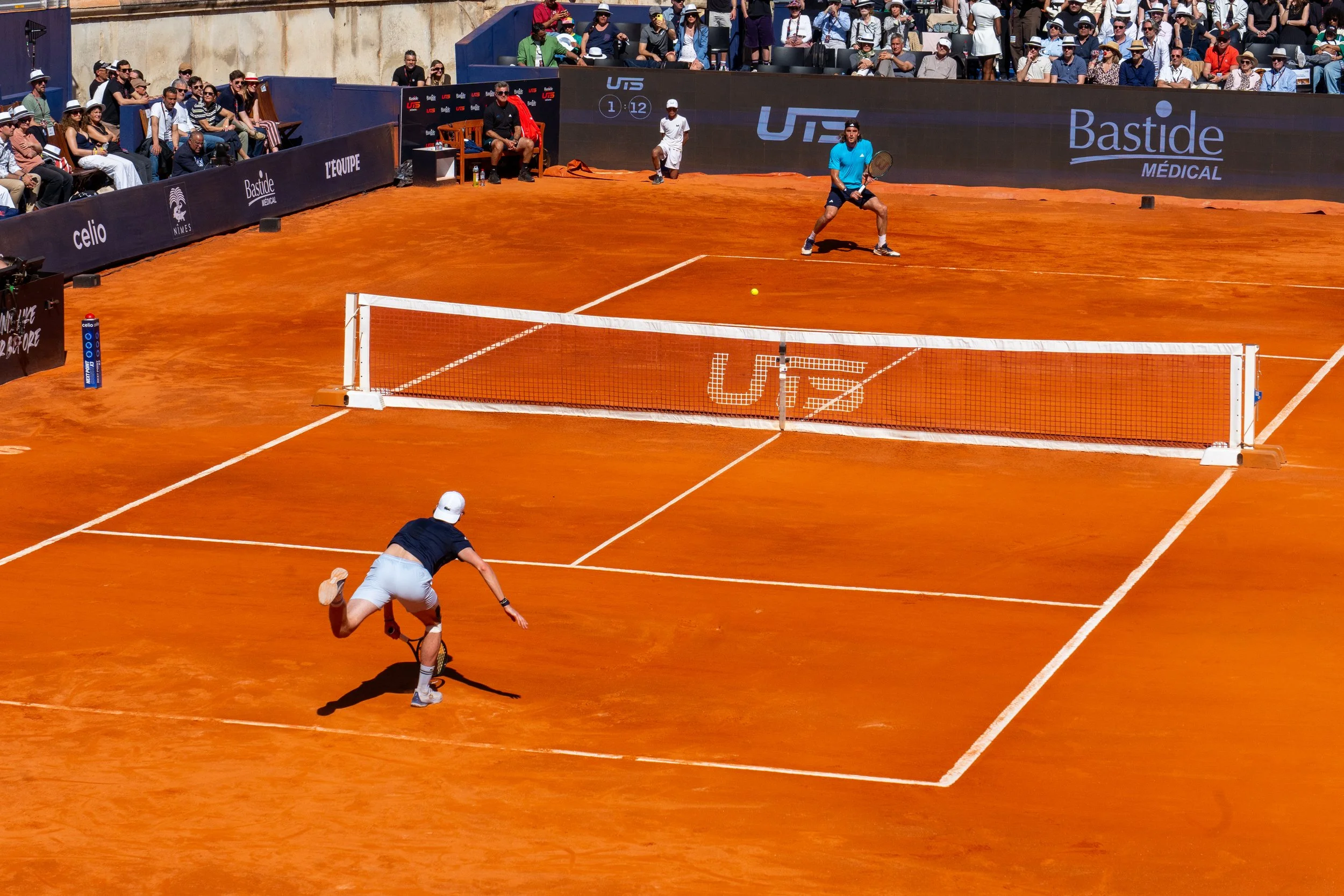 A tennis match on a clay court with two players, one at the net and the other preparing to hit the ball, surrounded by spectators in the stands.