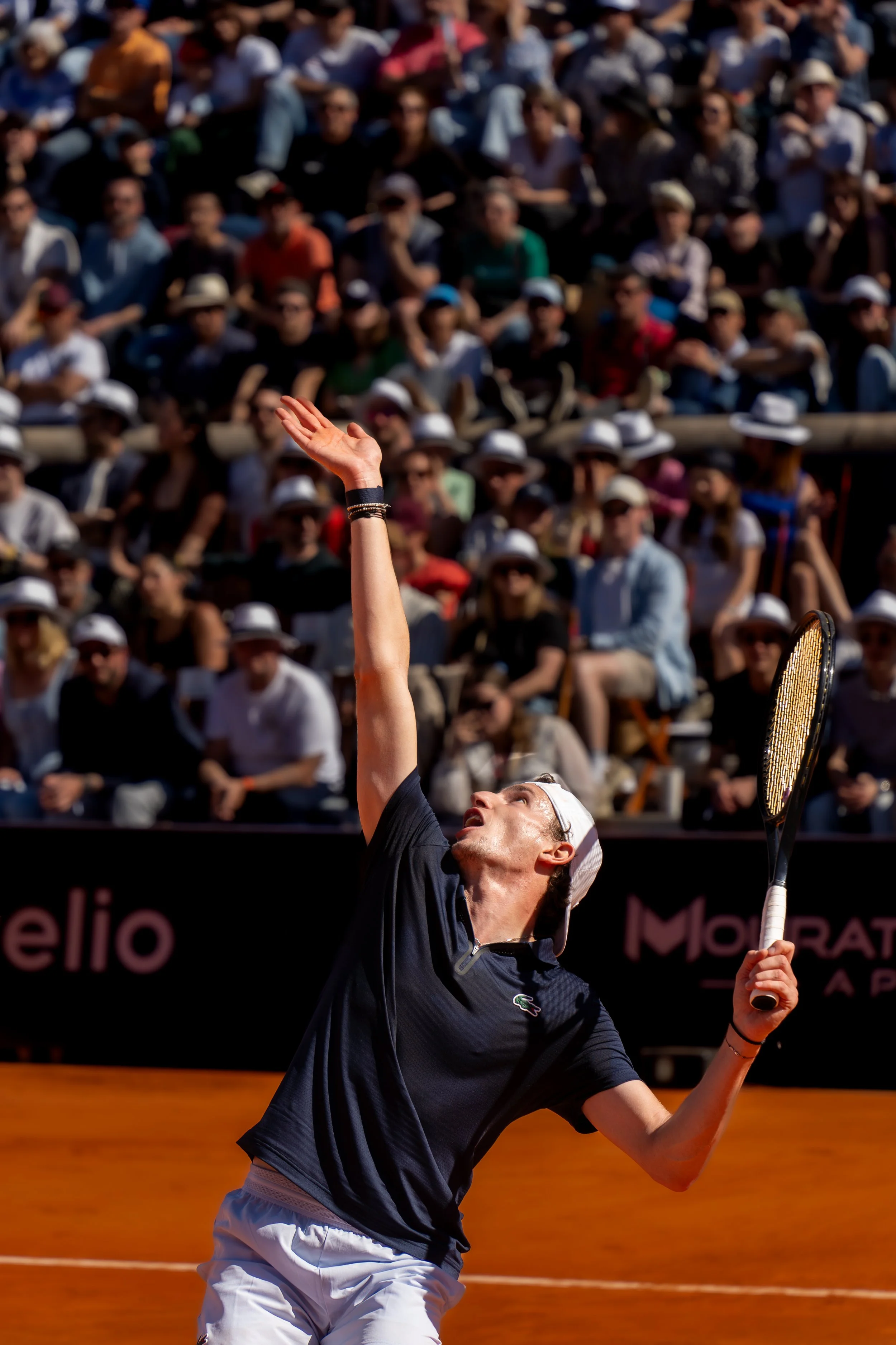 A male tennis player in a black shirt and white cap serving on a clay court during a match with a large crowd in the stands watching