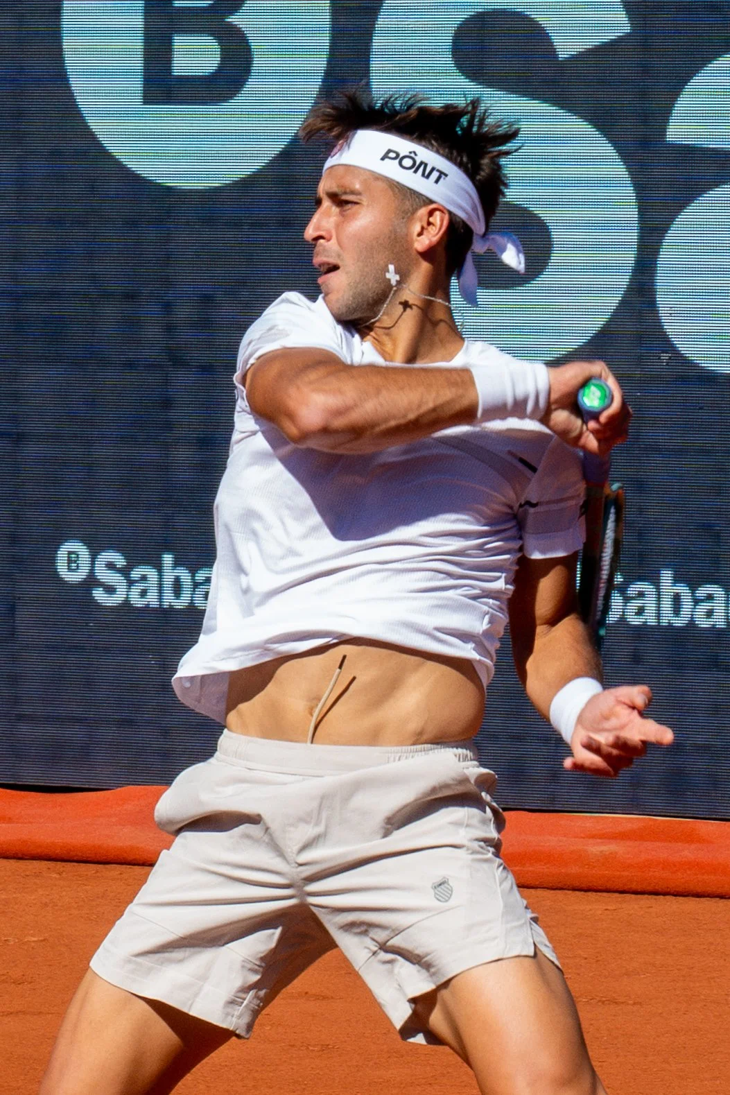 Tomas Etcheverry in white attire and headband is in the middle of a backhand stroke on a clay court, with a digital screen in the background.