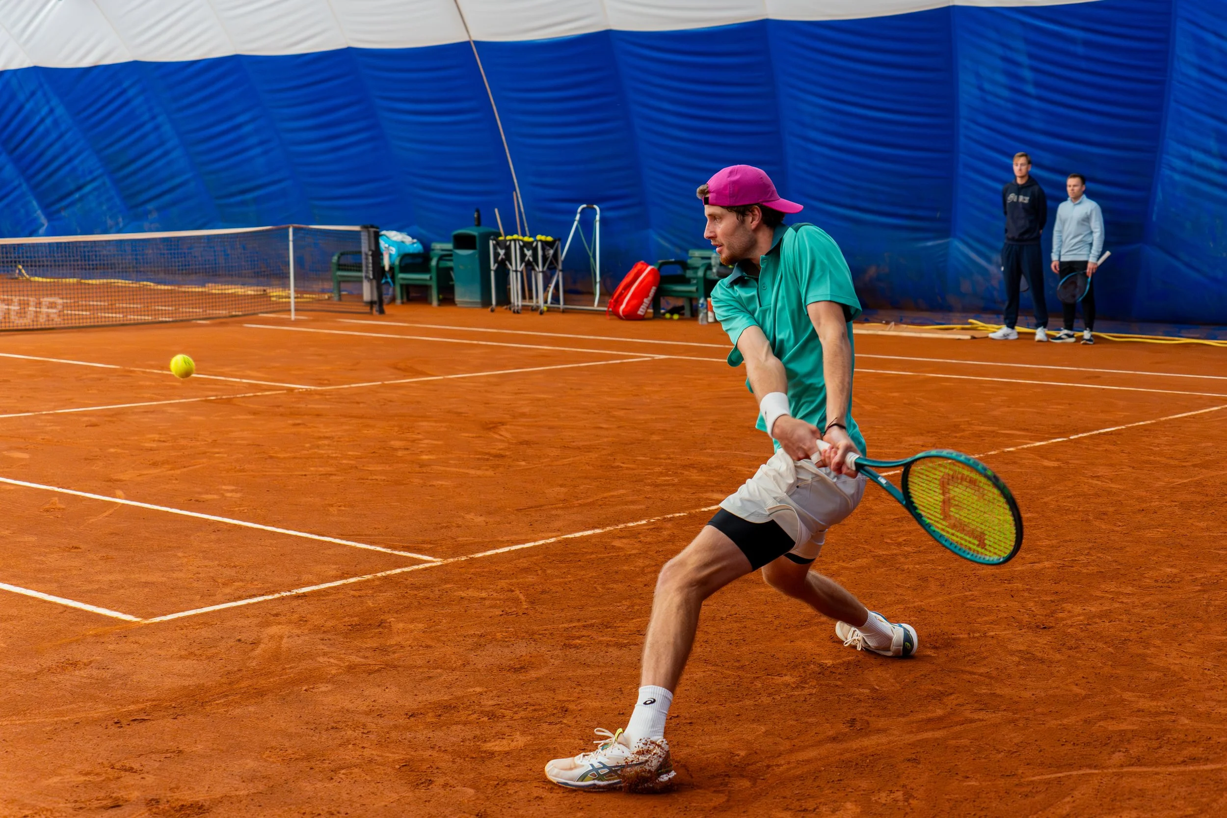A man in a green shirt and white shorts playing tennis on a clay court, preparing to hit a ball with his racket. Two people stand in the background near the blue inflatable dome.