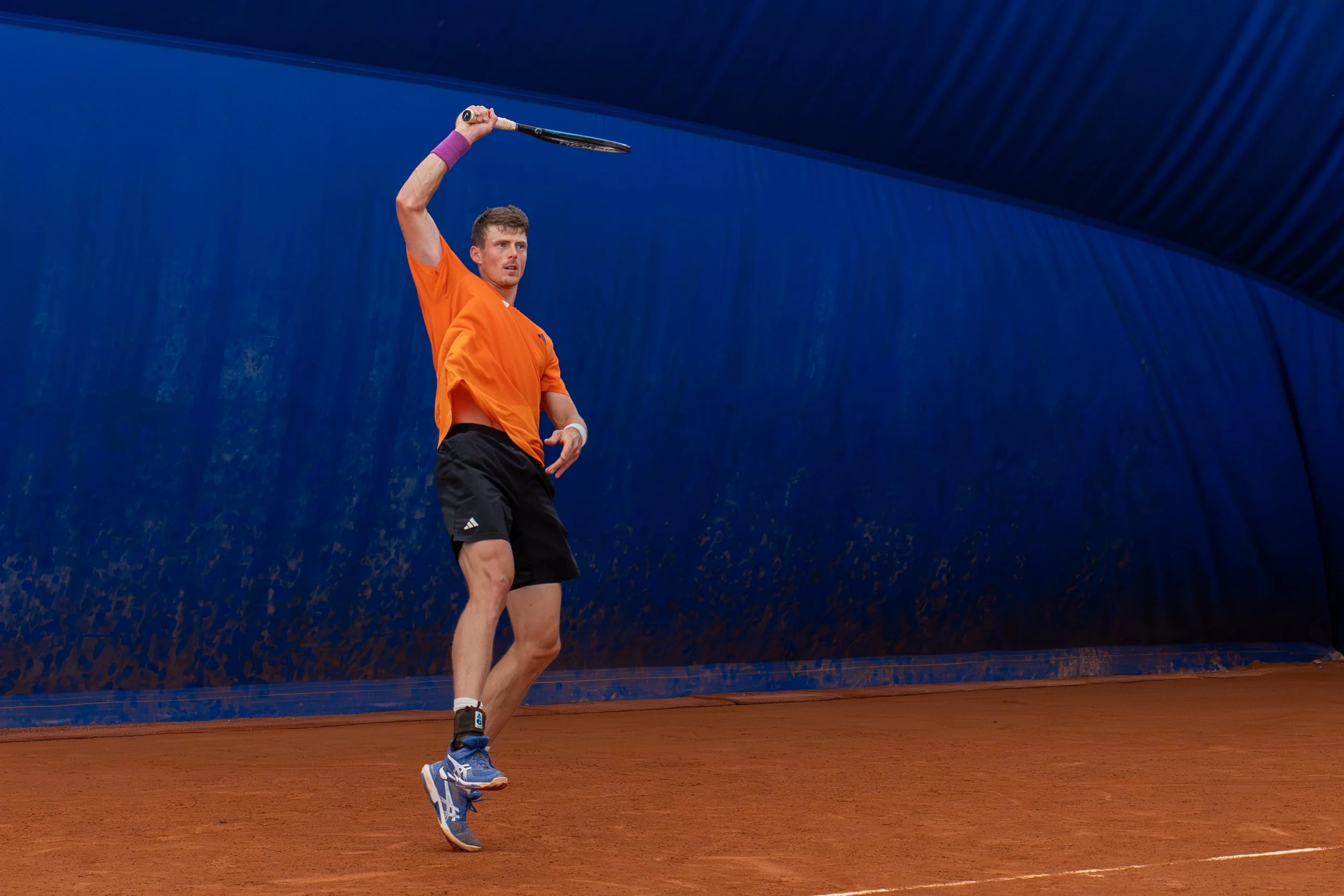 Tennis player in orange shirt and black shorts hitting a tennis ball with a racket on a clay court.
