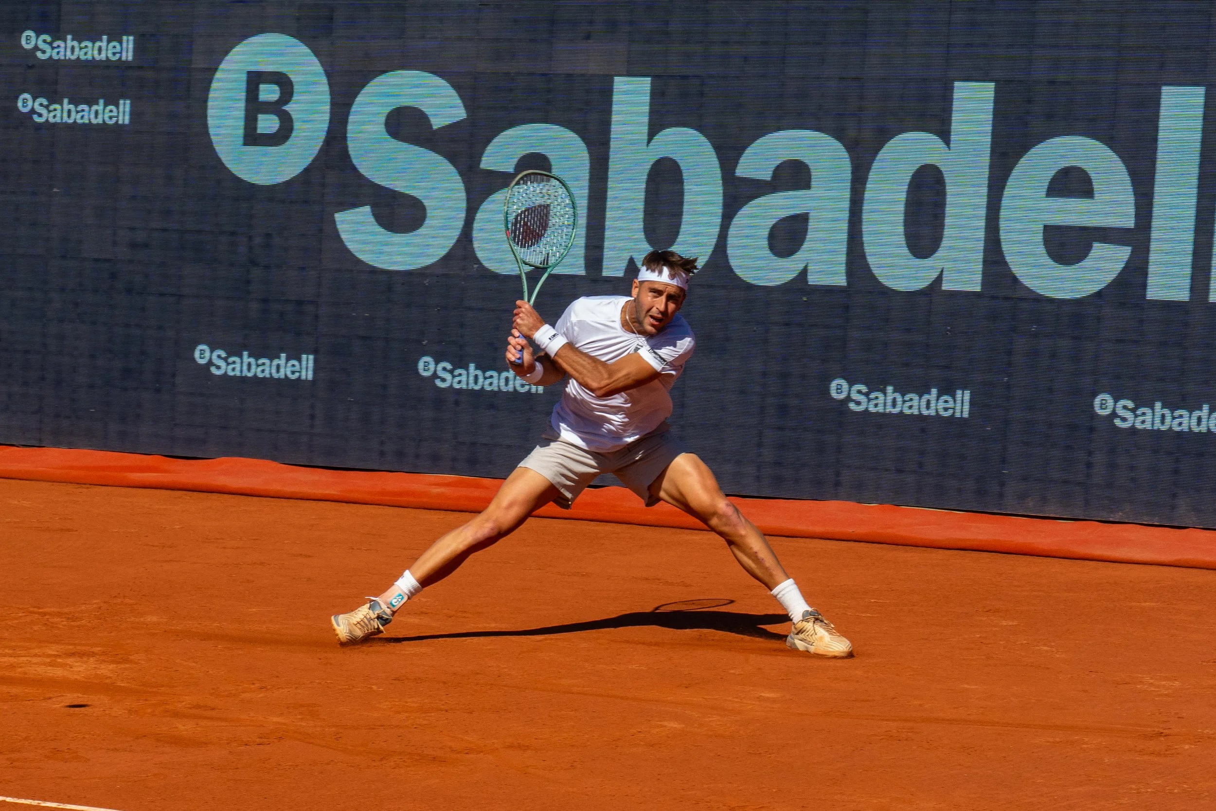 Tomas Etcheverry in a white shirt, beige shorts, and sneakers is lunging to hit a tennis ball with his racket on a clay court. There is a large electronic sign in the background with the word 'Sabadell' displayed.