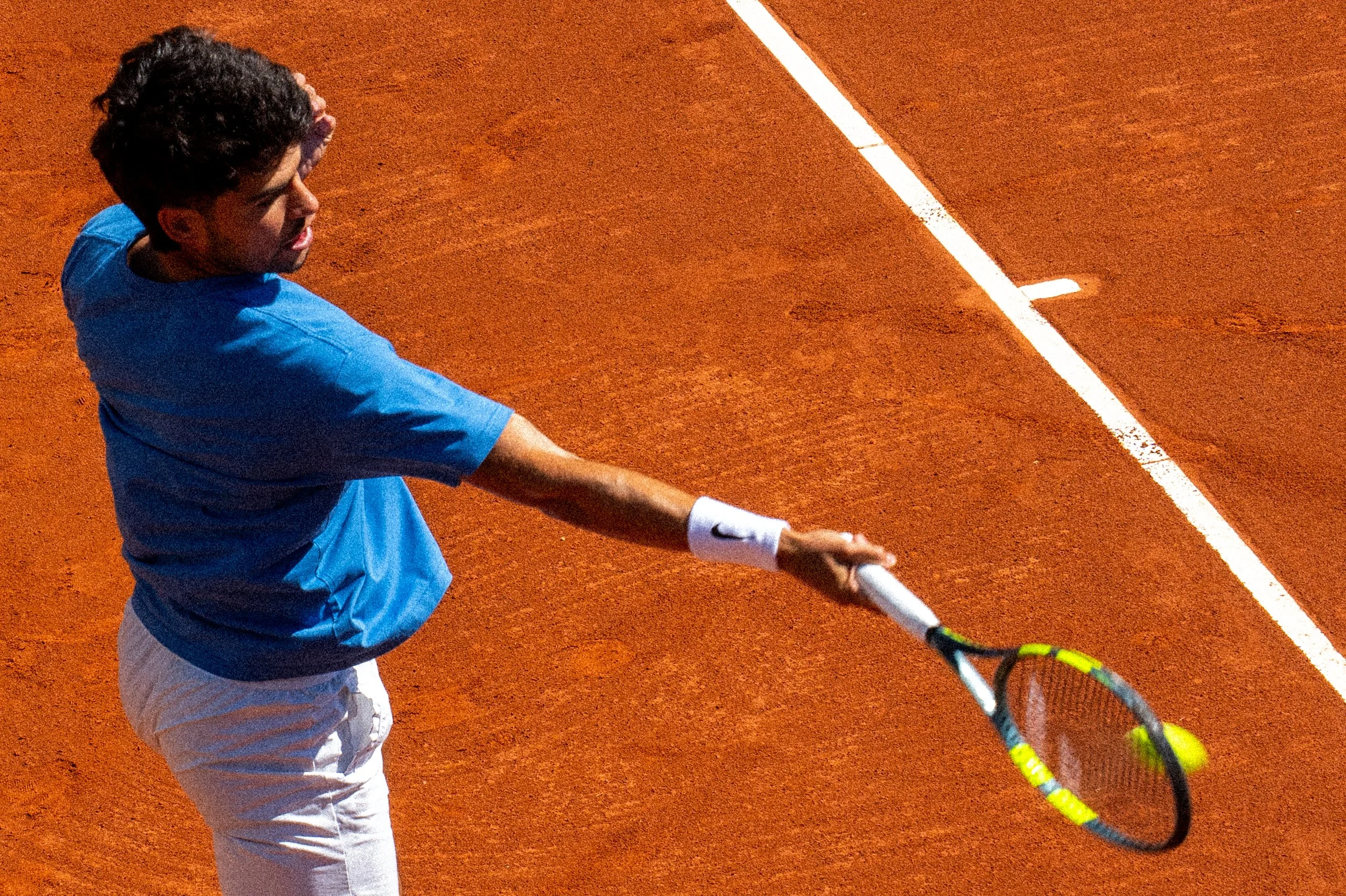 Tennis player Carlos Alcaraz preparing to hit a tennis ball on a red clay court at night.
