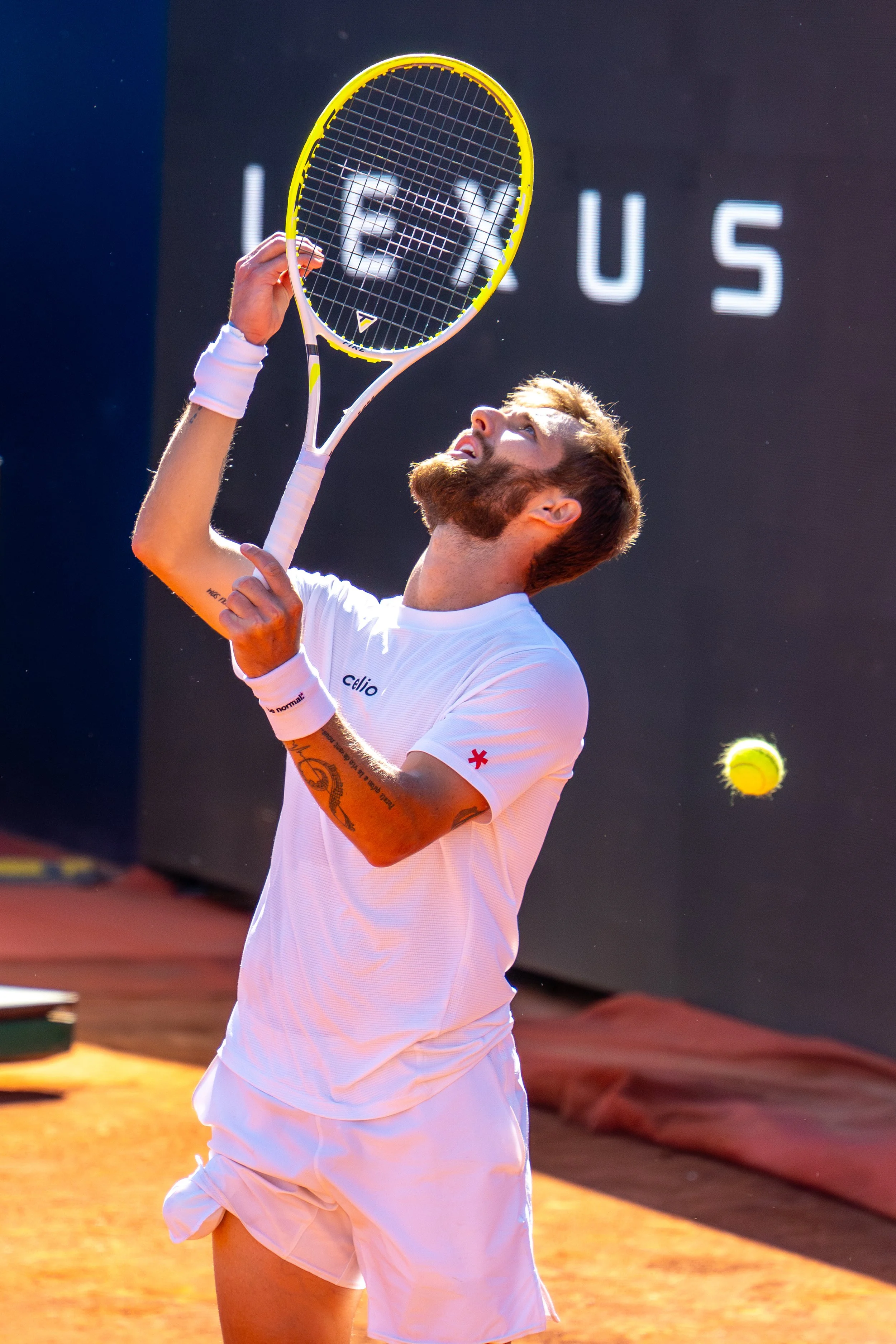 Corentin Moutet, dressed in a white shirt and shorts, prepares to serve on an outdoor clay court. There is a large digital screen displaying the word 'FOCUS' in the background, and a tennis ball is in midair near him.