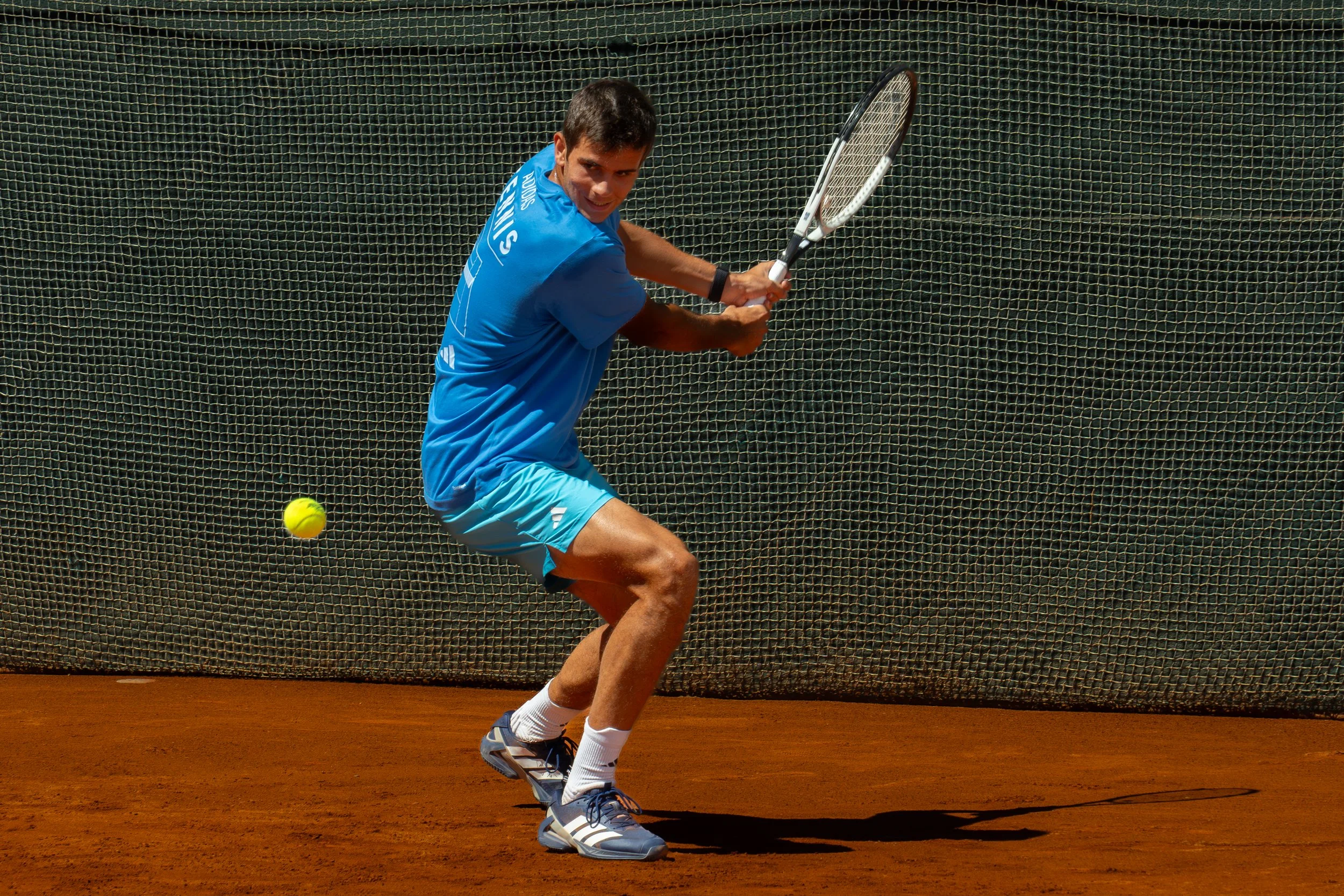 Rafa Jodar in a blue shirt and shorts preparing to hit a tennis ball on a clay court, with a net in the background.