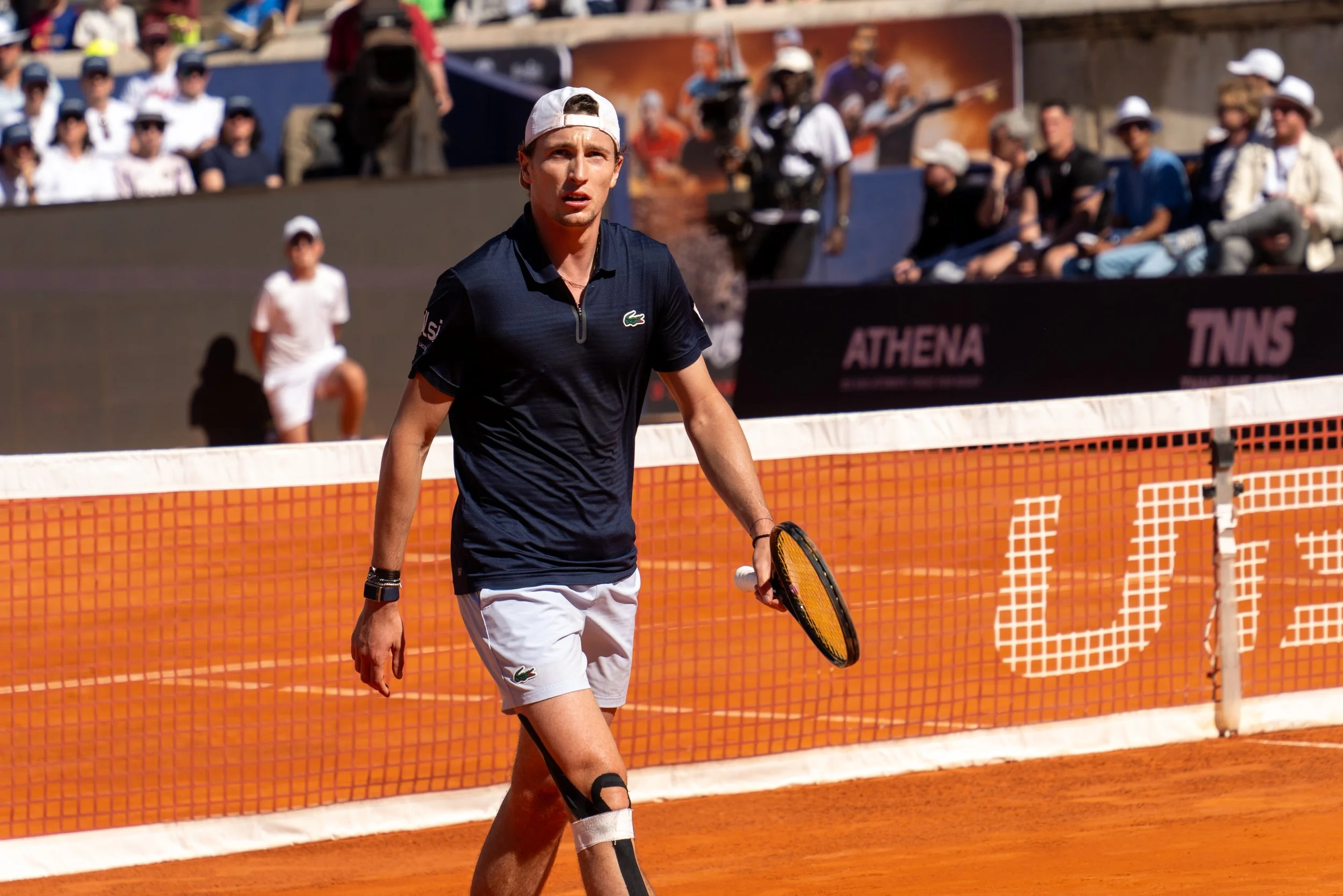 A male tennis player on a clay court, holding a tennis racket, wearing a navy shirt, white shorts, and a white cap, with an audience watching in the background.