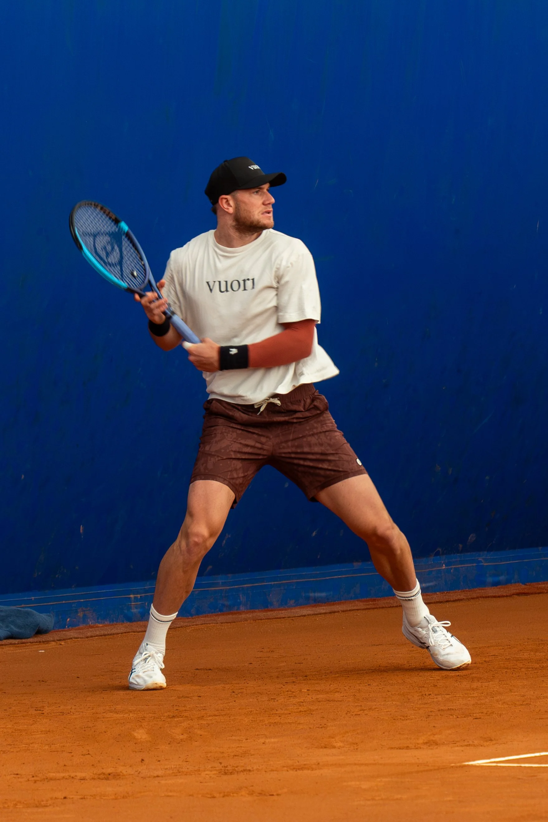 A man playing tennis on a clay court against a blue wall. He is wearing a white t-shirt, brown shorts, white tennis shoes, and a black cap, holding a tennis racket.