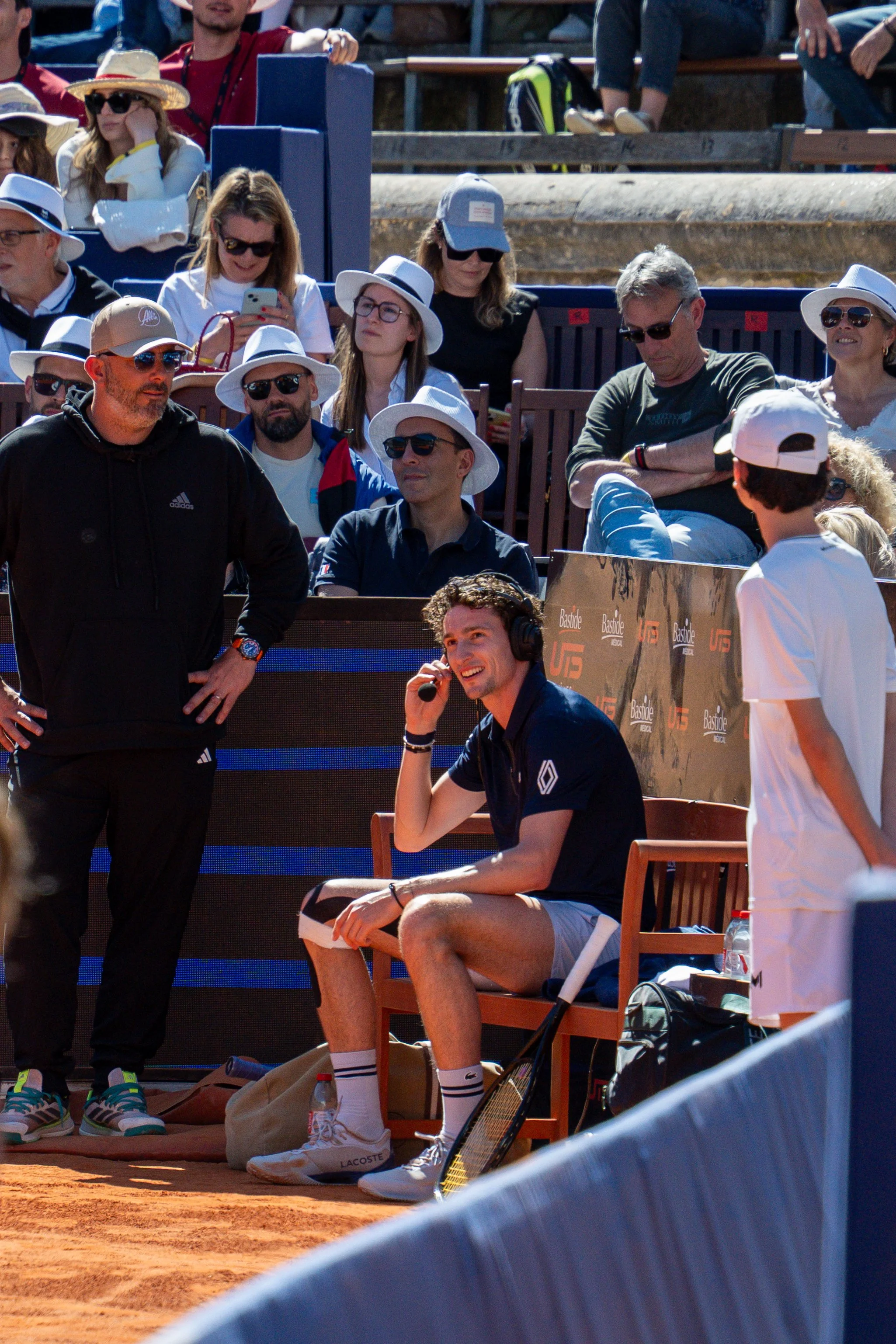 A tennis player sitting on a bench, speaking into a headset, surrounded by coaches and spectators at an outdoor match.