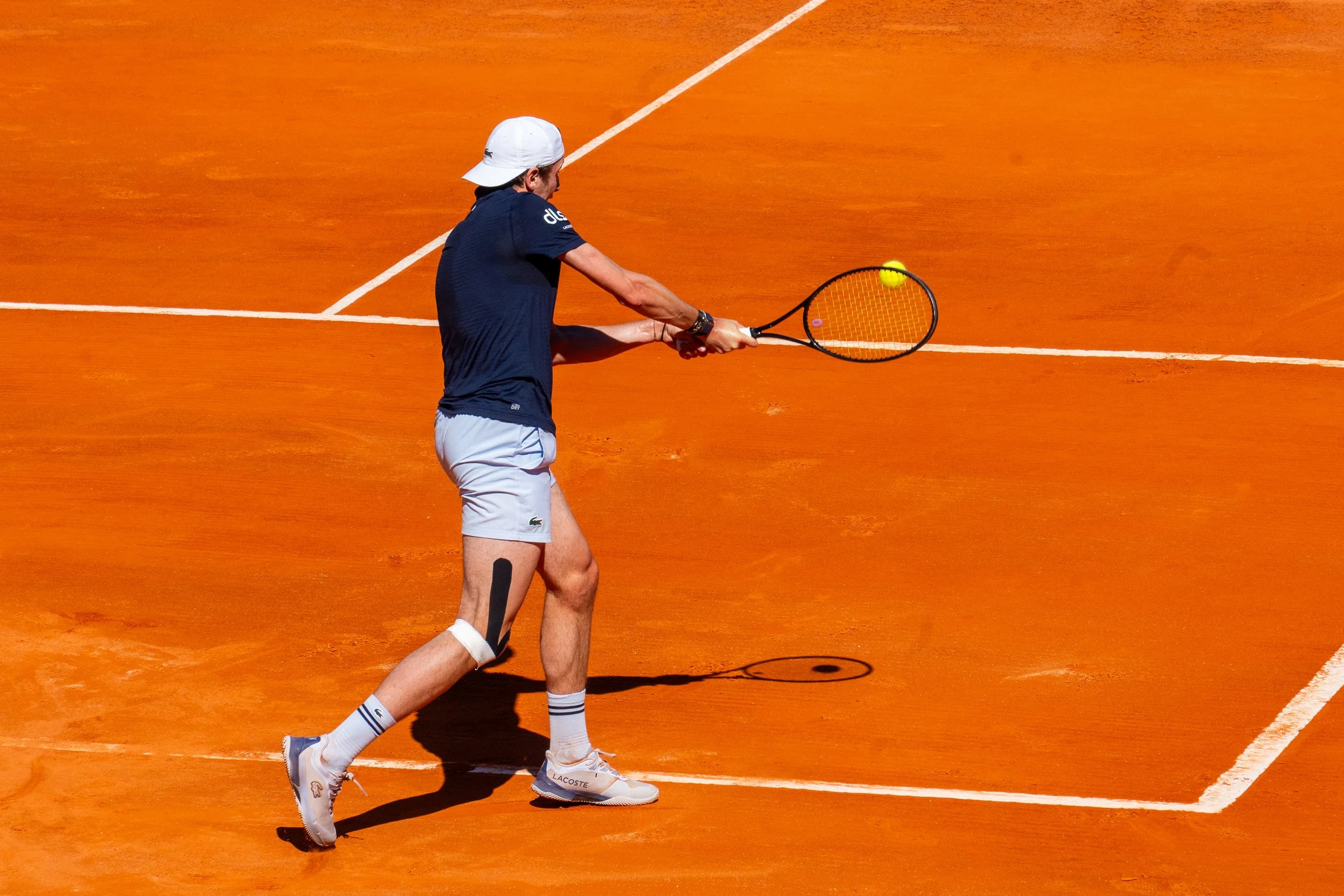 A man in a white cap, navy blue shirt, and white shorts on a tennis court hitting a tennis ball with a racquet.