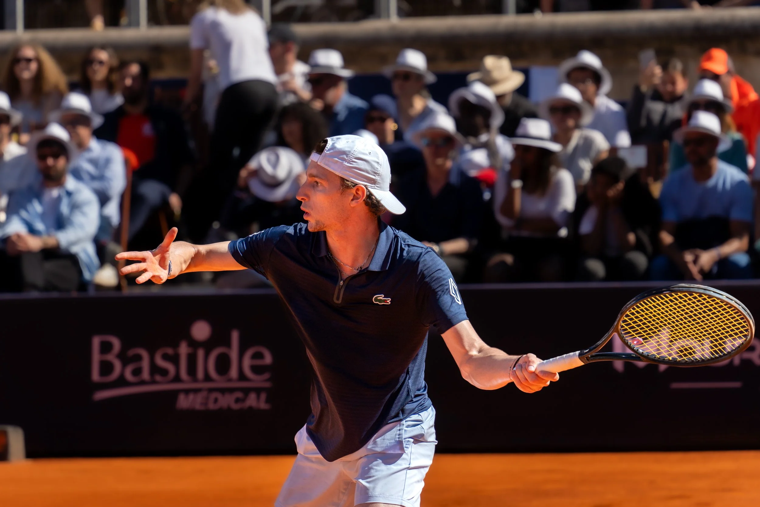 A male tennis player in a navy blue shirt and white shorts is hitting a forehand shot during a match on a clay court, with spectators wearing hats and sunglasses in the background.