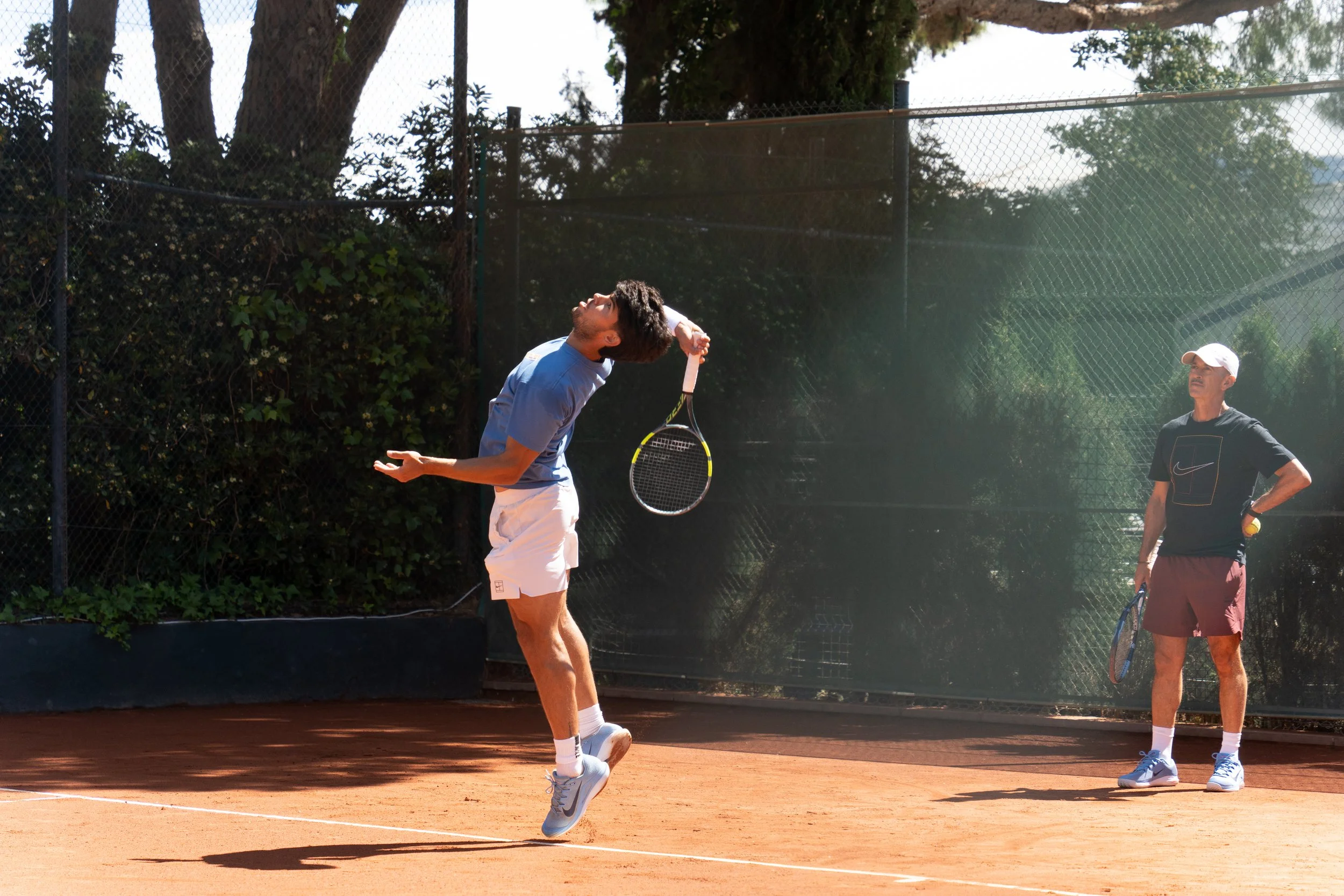 Carlitos Alcaraz, from Murcia, Spain, playing tennis on a clay court, hitting a tennis ball with a racket in the air while a coach or companion watches nearby. The court is outdoors with trees and a fence in the background, and sunlight is shining.