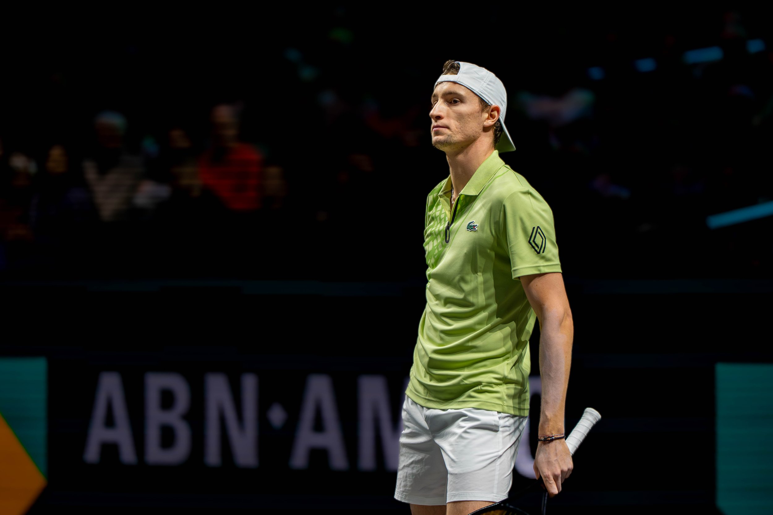A male tennis player wearing a lime green shirt, white shorts, and a white cap, standing on a tennis court holding a racket with a serious expression.