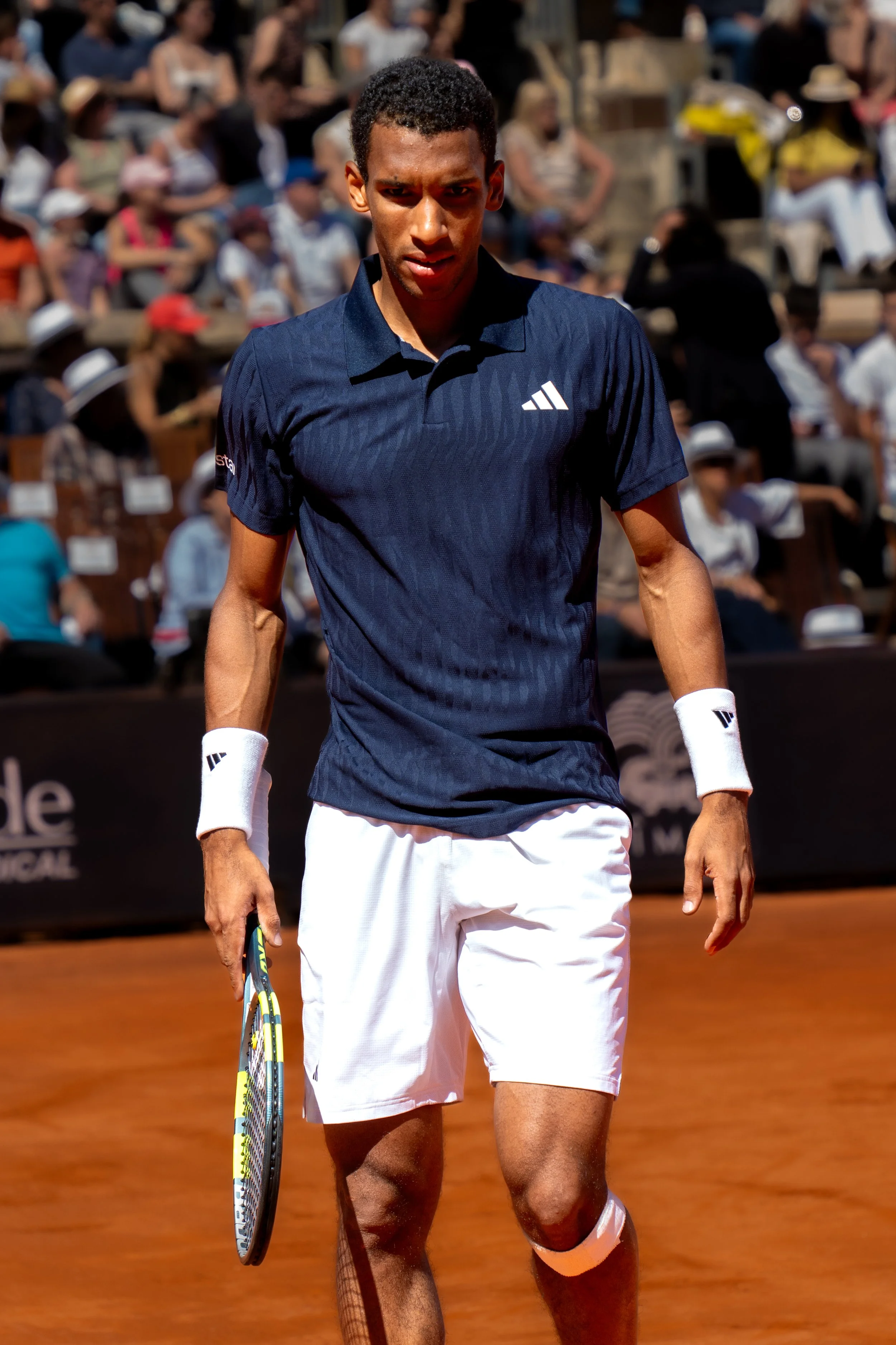 Male tennis player on clay court during match, wearing navy shirt and white shorts, holding tennis racket, with spectators in background.