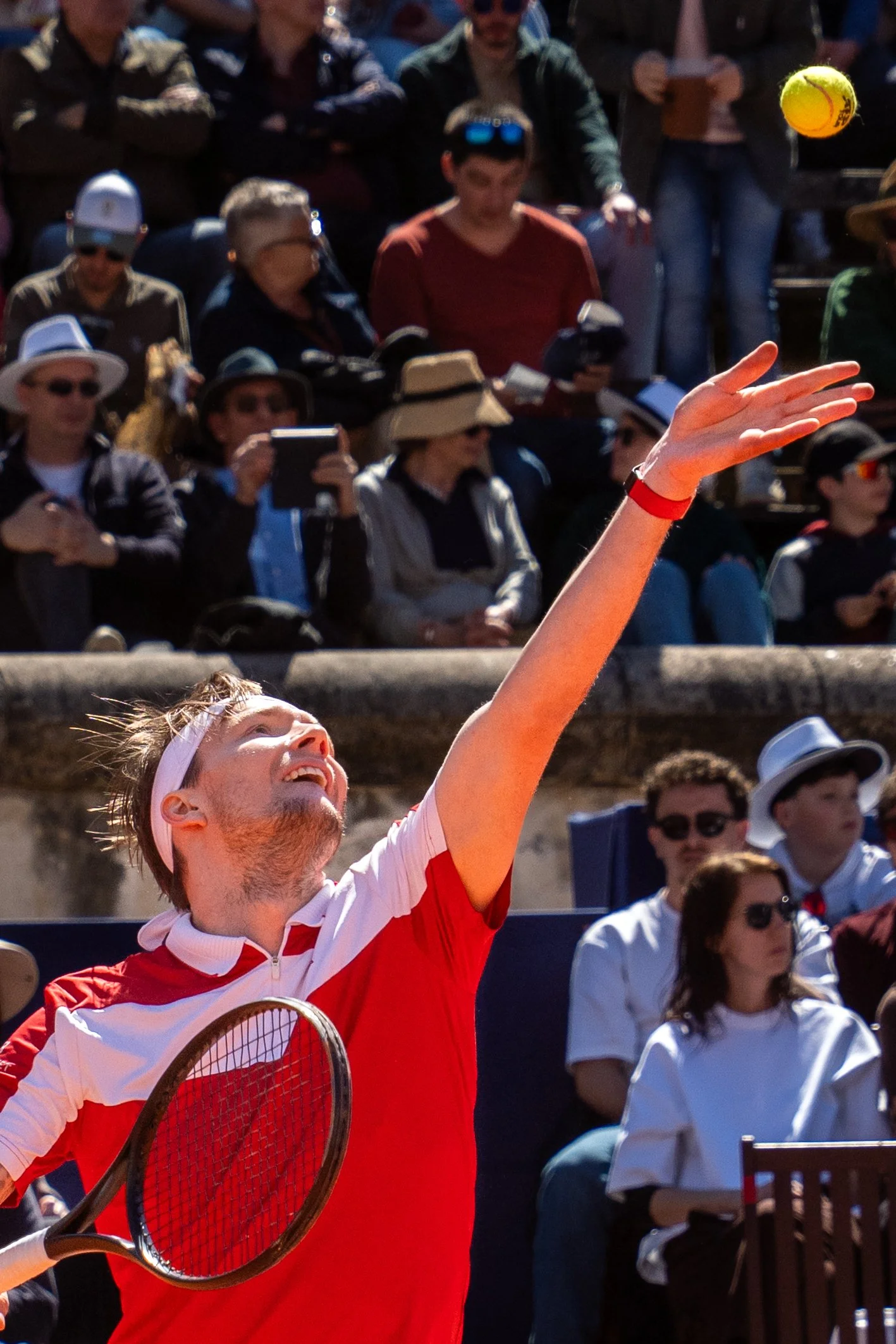 Tennis player in a red shirt hitting a ball during a match with spectators watching in the background.
