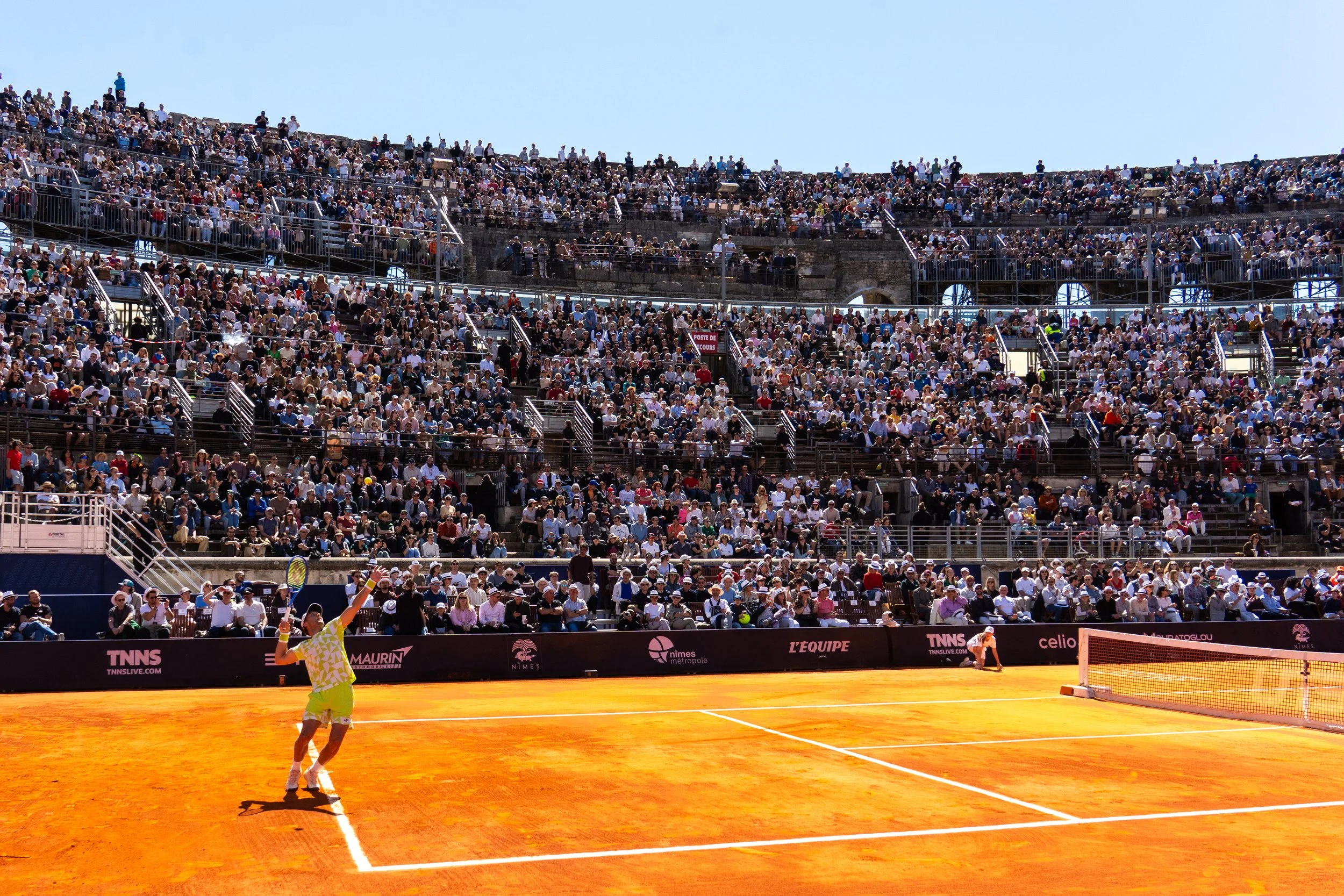 Tennis match at a large outdoor stadium with a packed audience, a player in a yellow outfit preparing to serve, on a clay court under a clear sky.