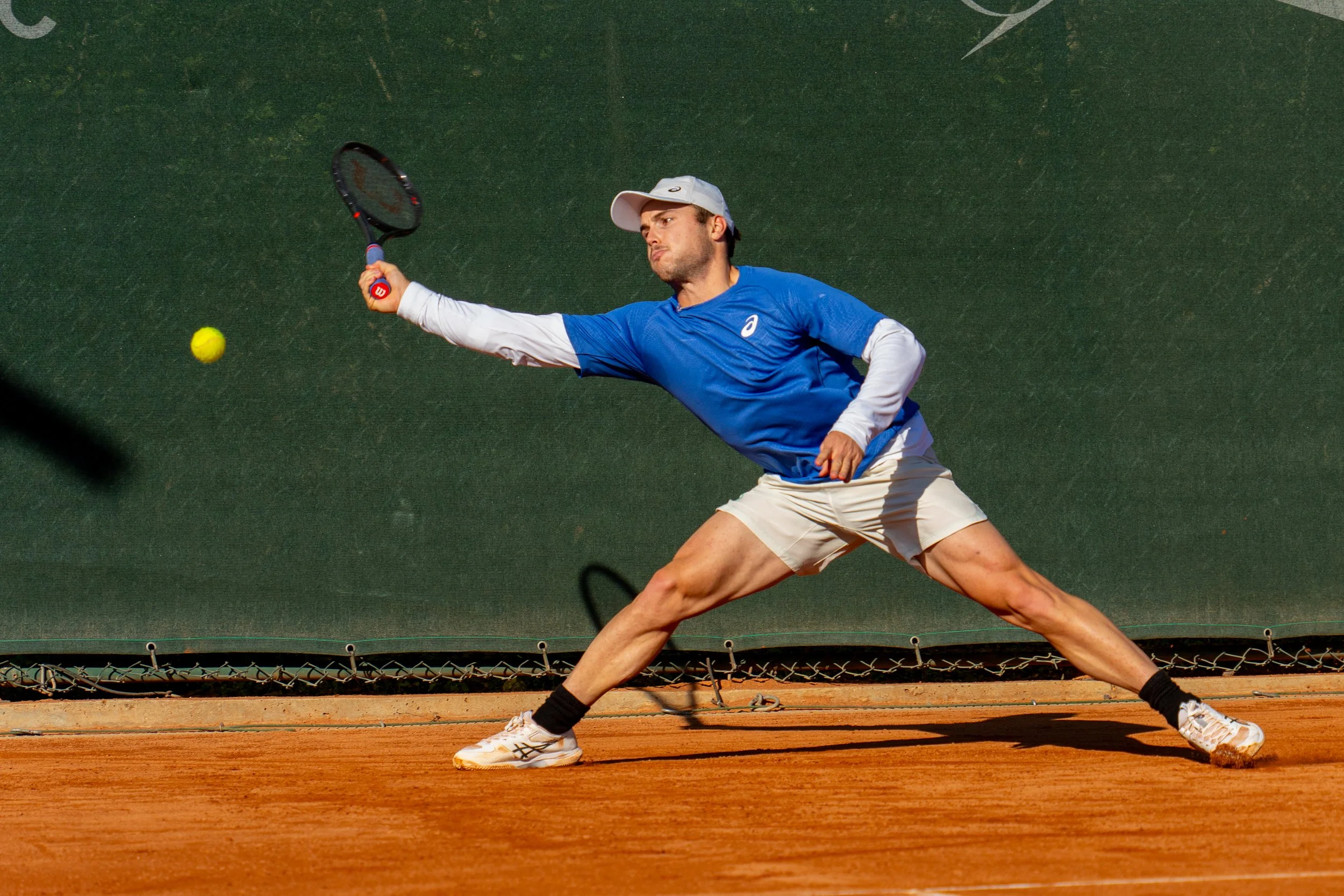A man in a blue shirt and white shorts playing tennis on a clay court, reaching out with his racket to hit a yellow tennis ball.