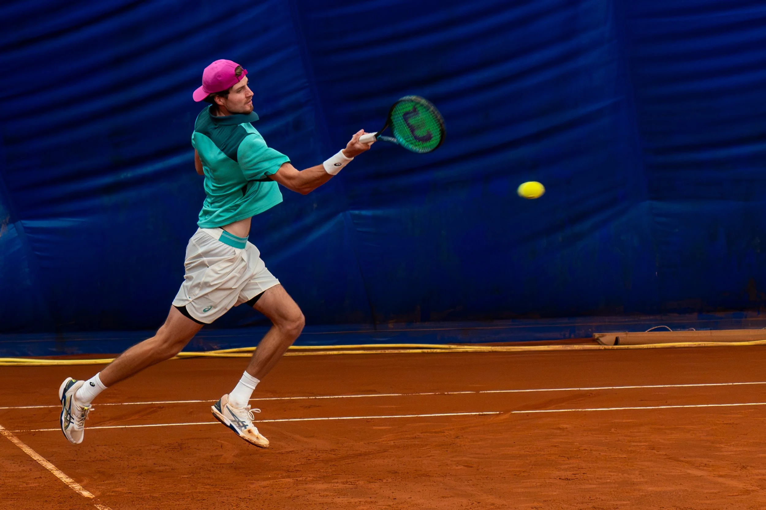 A man playing tennis on a clay court, wearing a teal shirt, white shorts, a pink cap worn backwards, and white sneakers, preparing to hit a yellow tennis ball with a green and black racket.