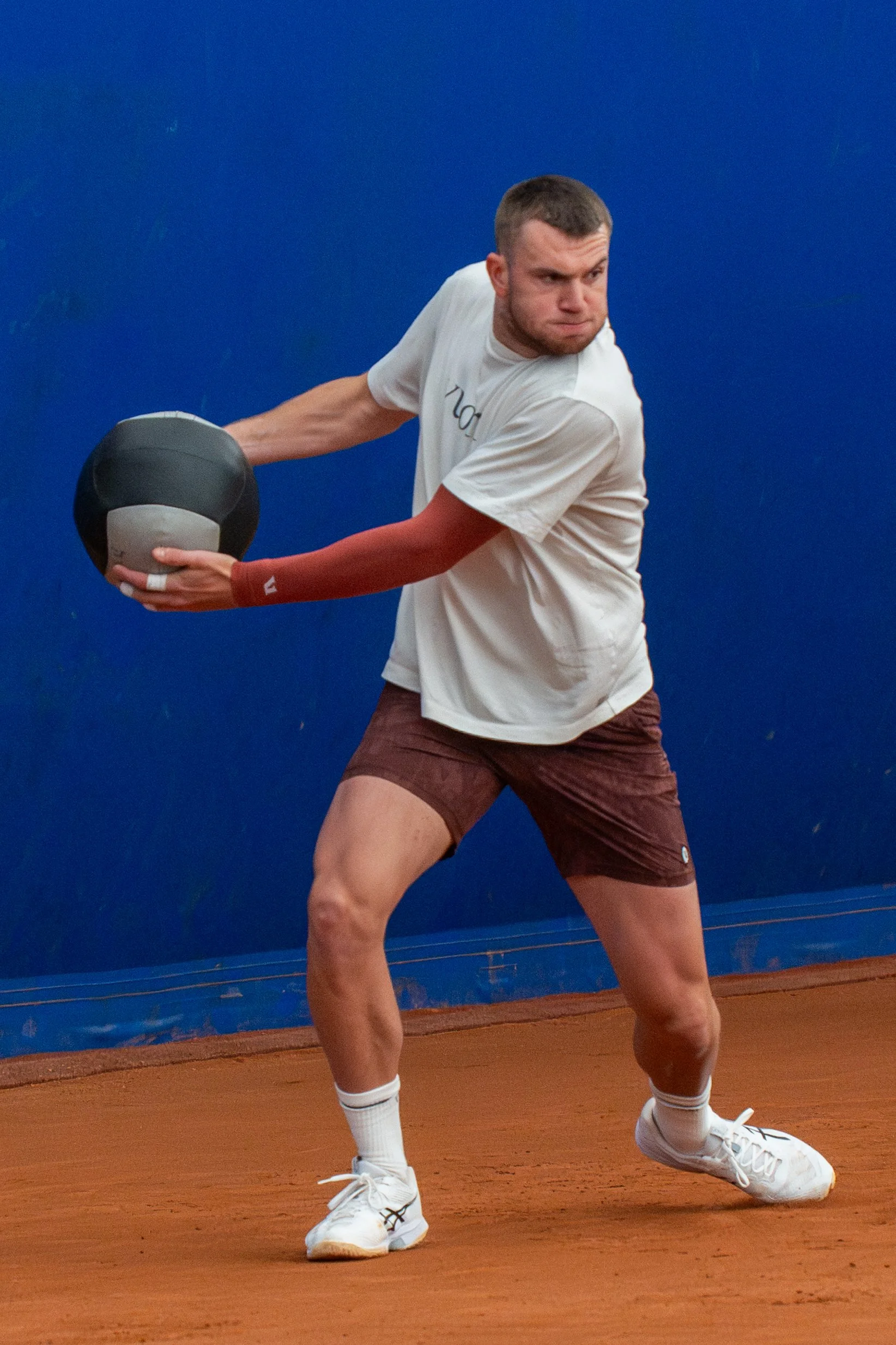 A male tennis player prepares to hit a tennis ball with a black medicine ball on a clay court, with a blue wall in the background.