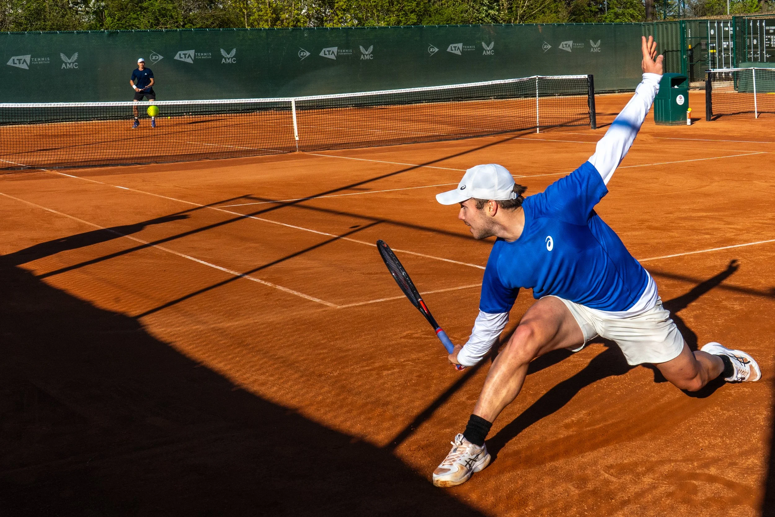 A male tennis player in a blue shirt and white shorts lunges to reach a tennis ball on a clay court, with another player in the background on the opposite side of the net.