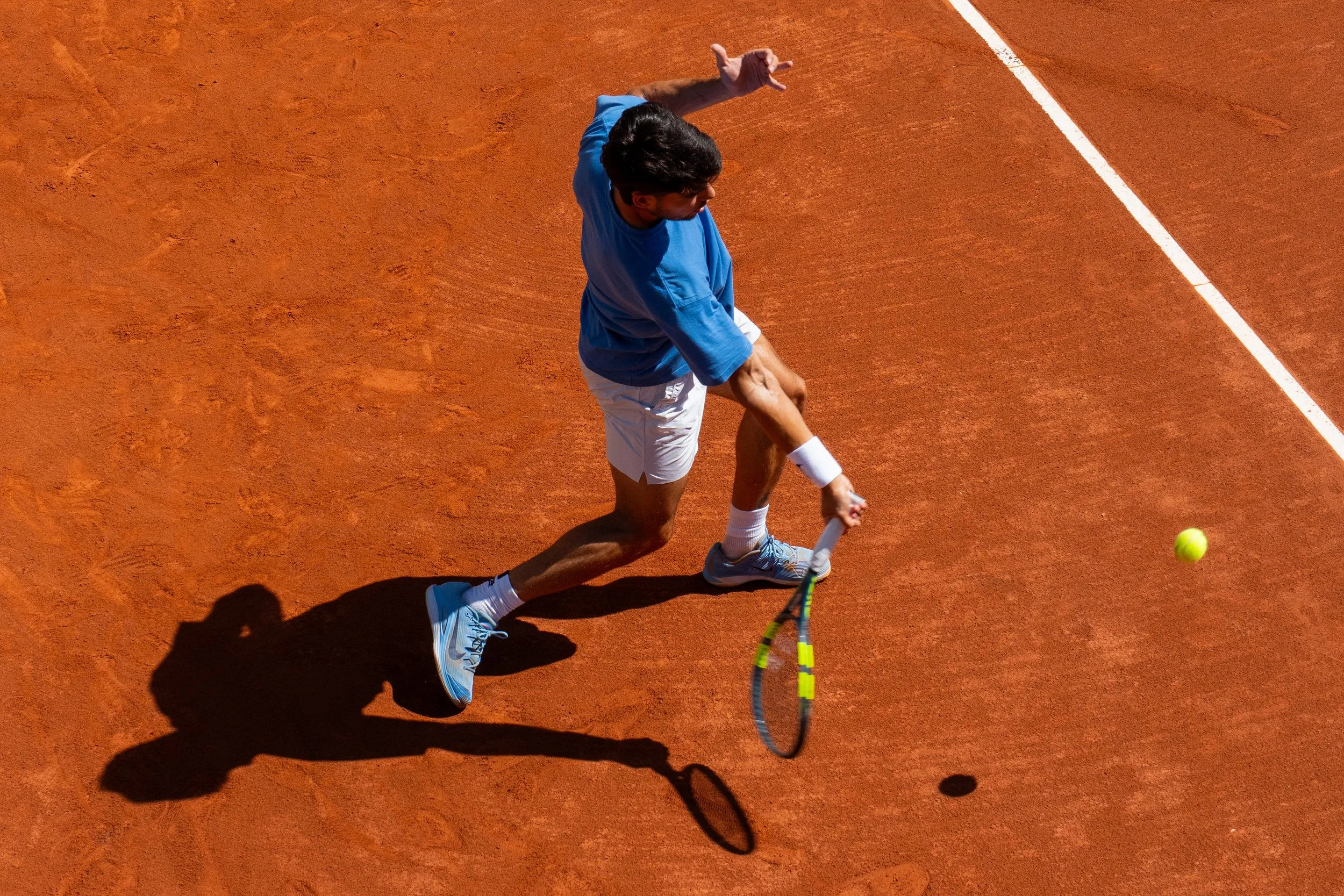 Tennis player Carlos Alcaraz on a clay court, about to hit a tennis ball with his racket. He is wearing a blue shirt, white shorts, and blue sneakers, with a shadow cast on the court.