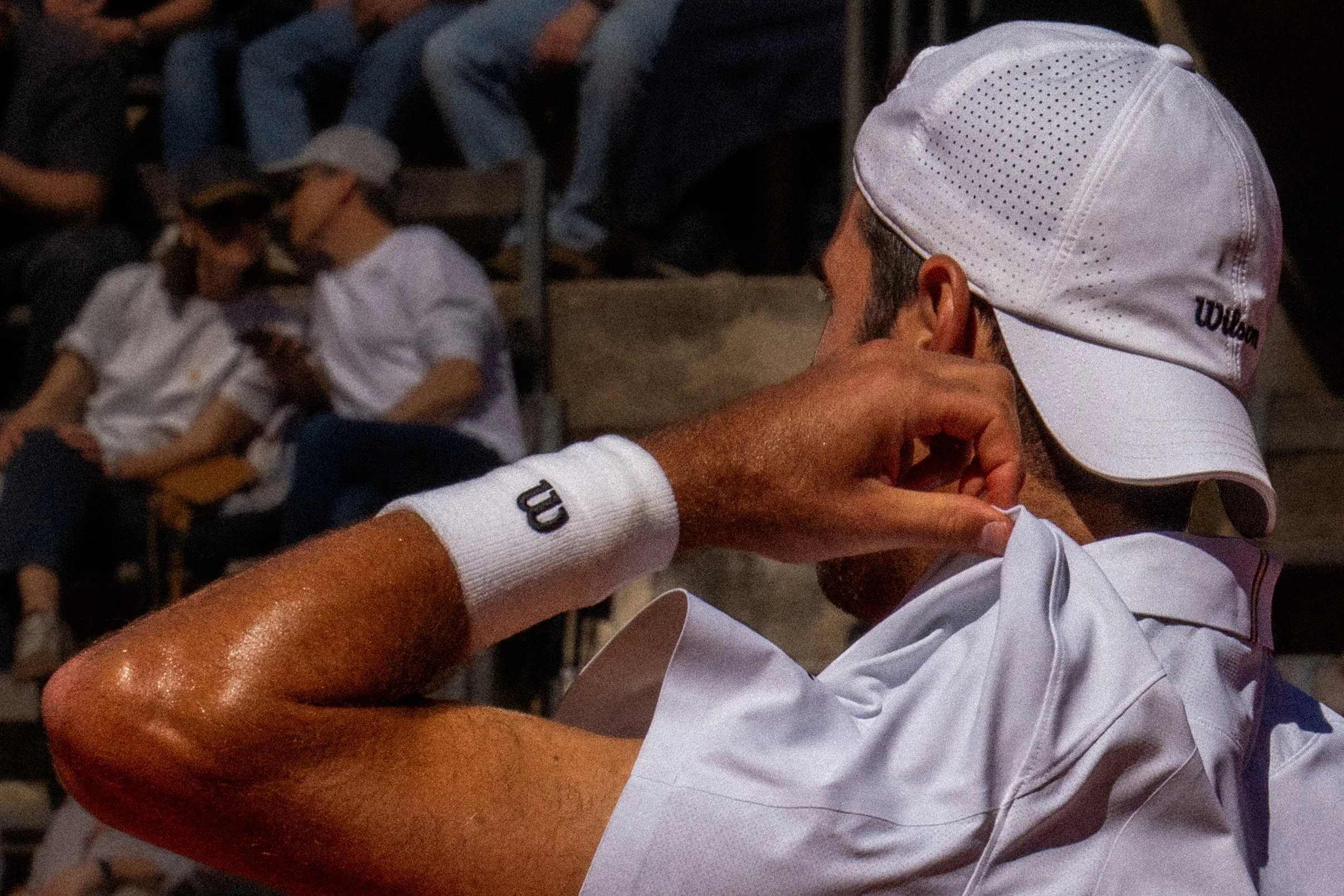 A close-up of a tennis player wearing a white Wilson cap and wristband, adjusting his ear during a match, with spectators sitting in the background.