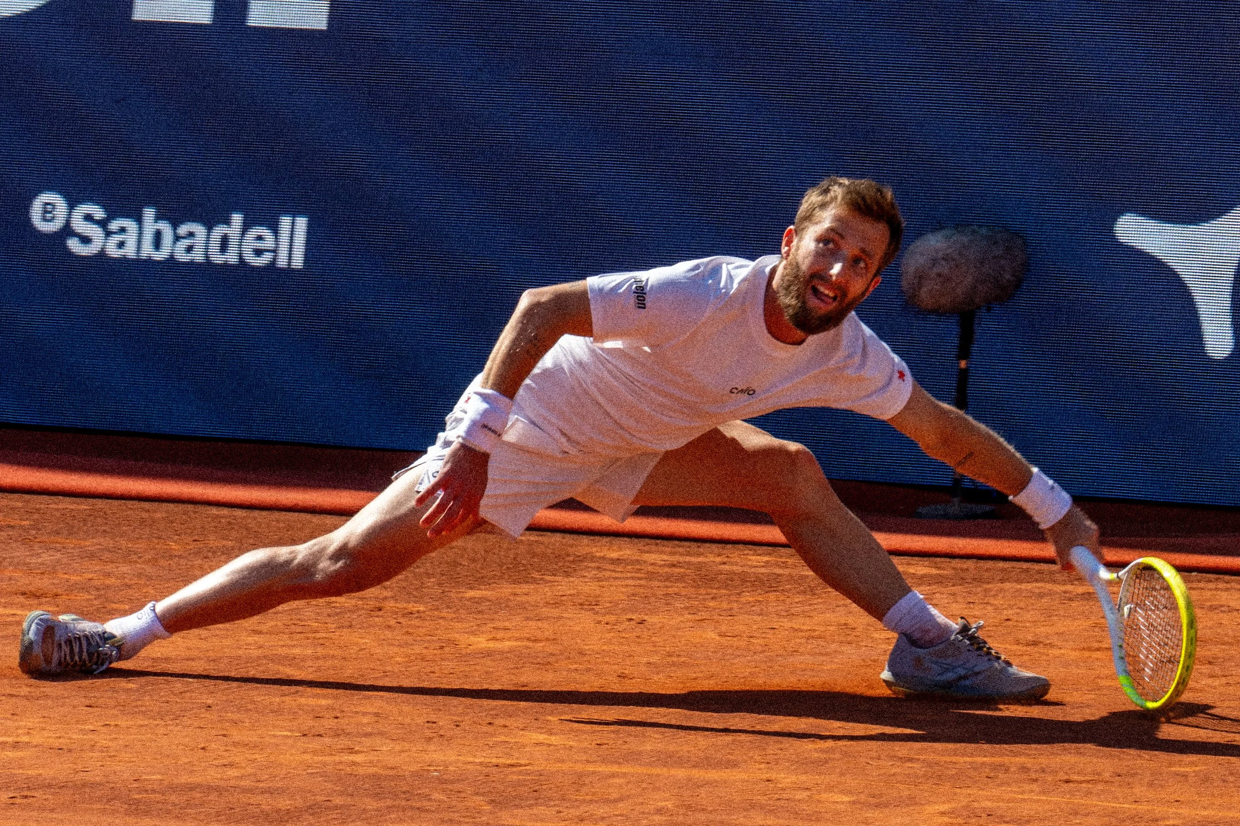A French tennis player Corentin Moutet in a white shirt and shorts stretching on a red clay tennis court in Barcelona, Spain, with a blue advertisement banner in the background.