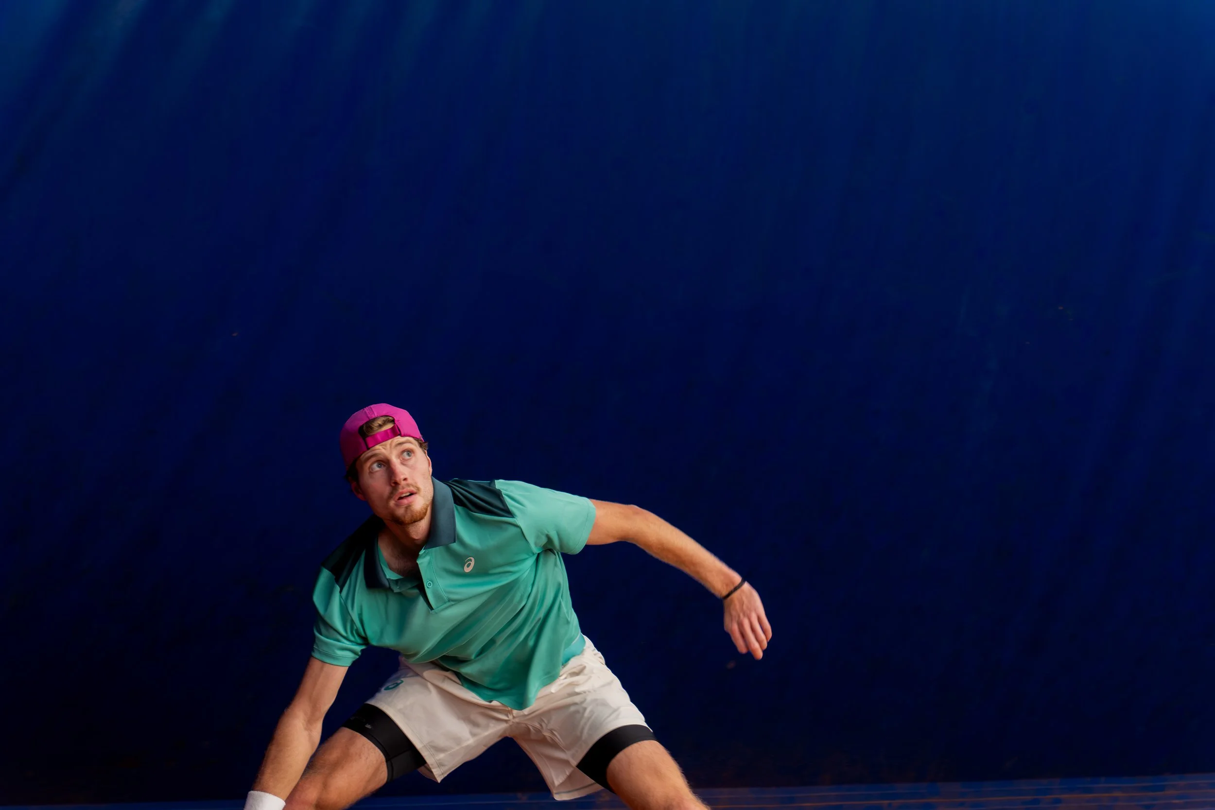 A man playing tennis, wearing a pink cap, green shirt, and white shorts, crouched low and looking upwards against a dark background.