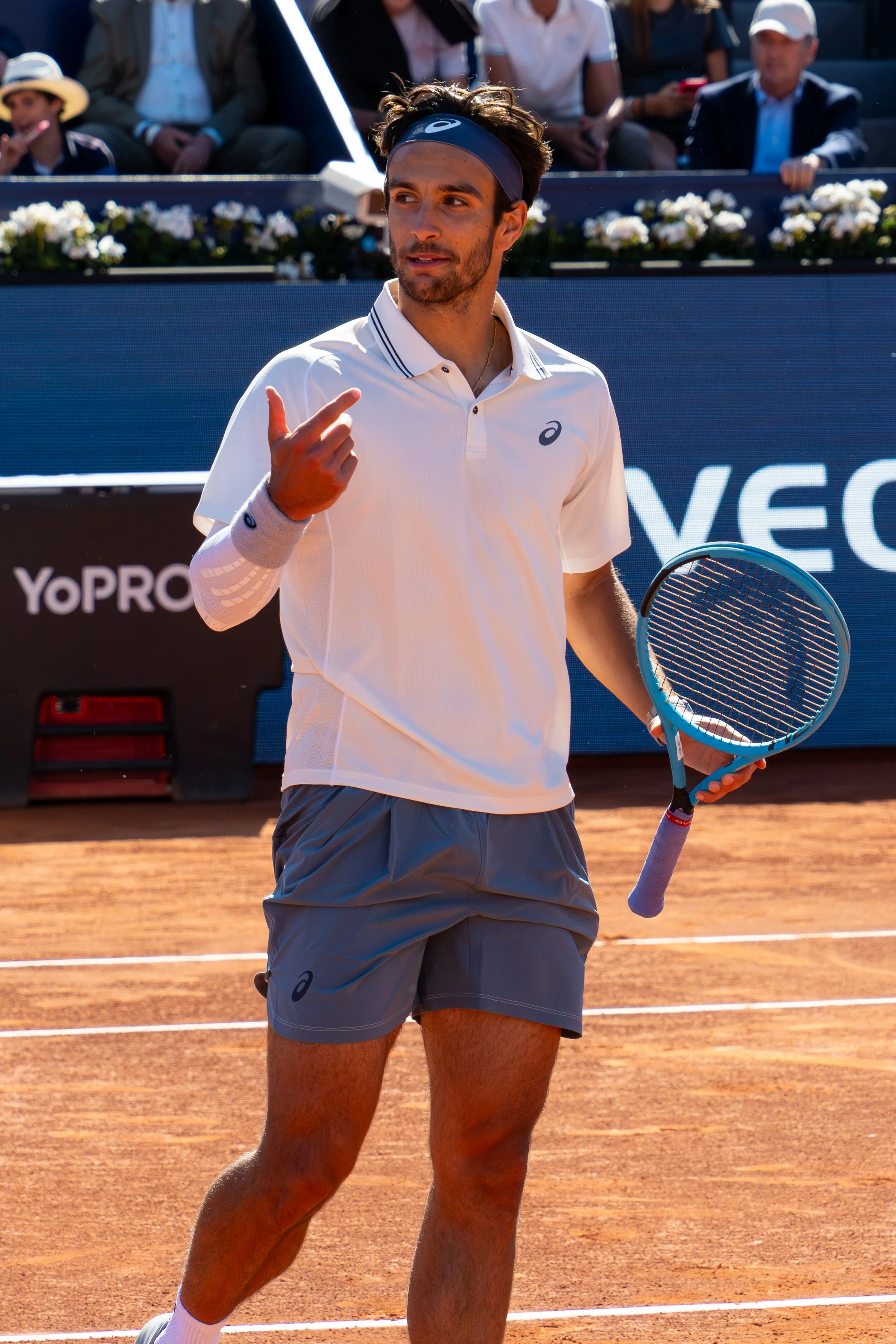 Lorenzo Musetti on a clay court, wearing a white shirt, gray shorts, a headband, and holding a tennis racket, gesturing with his hand during a match.