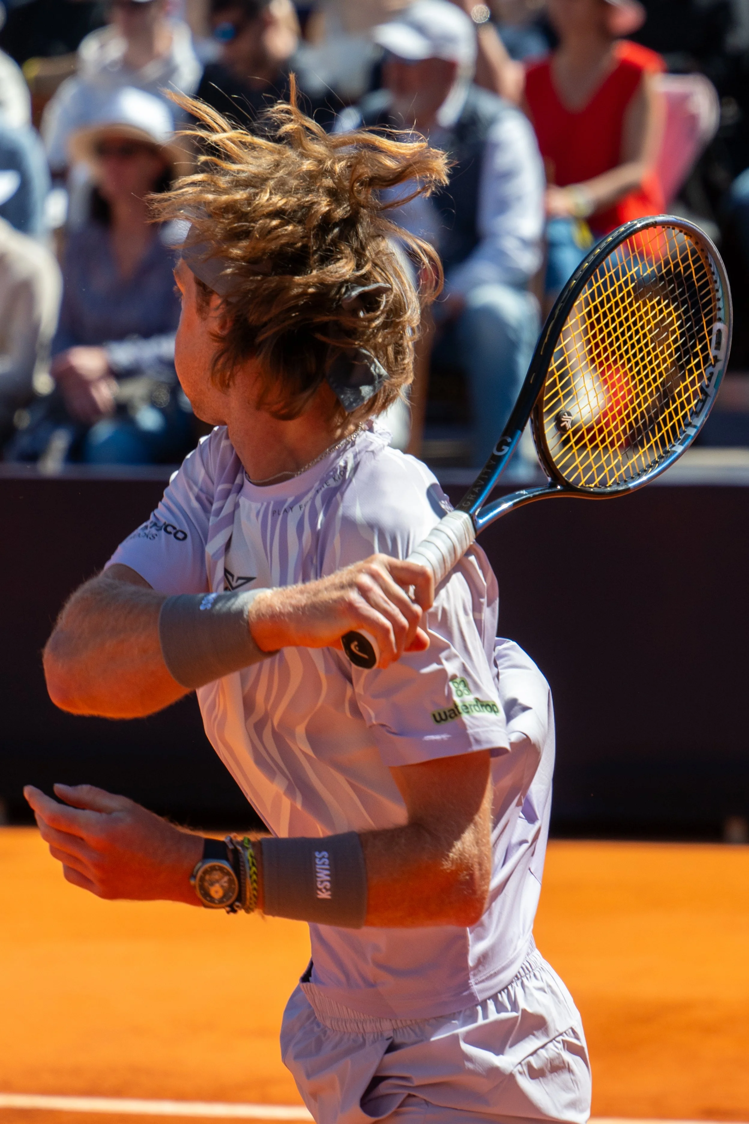 A tennis player with long, curly hair swings a tennis racket on a tennis court, with spectators watching in the background.