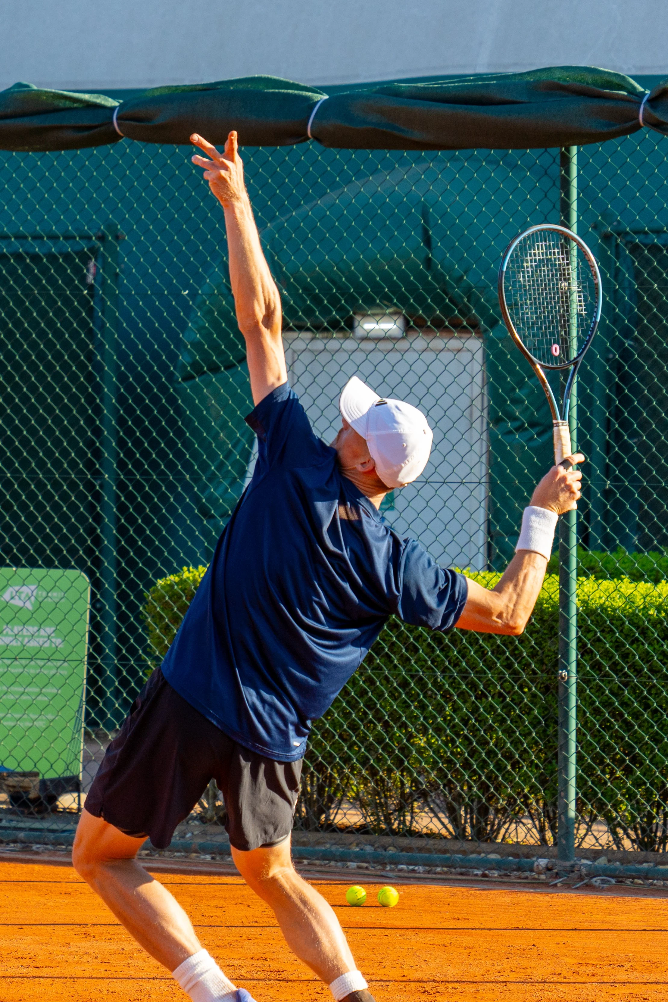 Male tennis player preparing to hit a serve on a clay court, wearing a white cap, dark blue shirt, black shorts, and white wristbands.