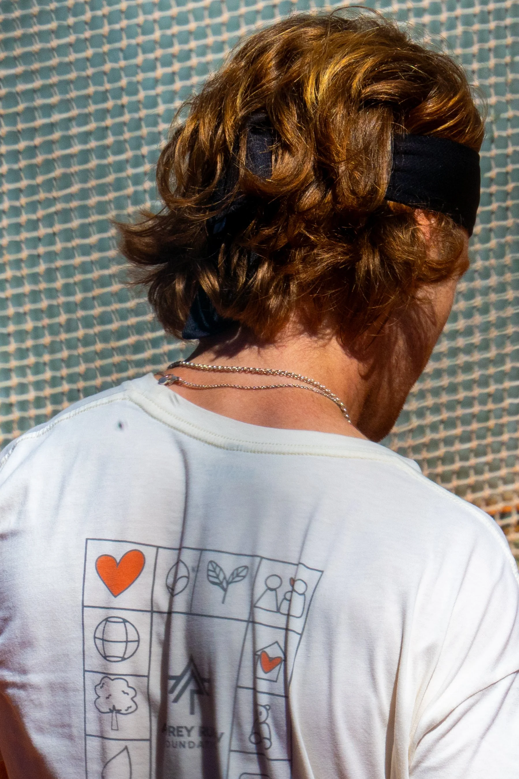 Back view of Russian tennis player Andrey Rublev wearing a black headband and a white T-shirt with a graphic design on the front.