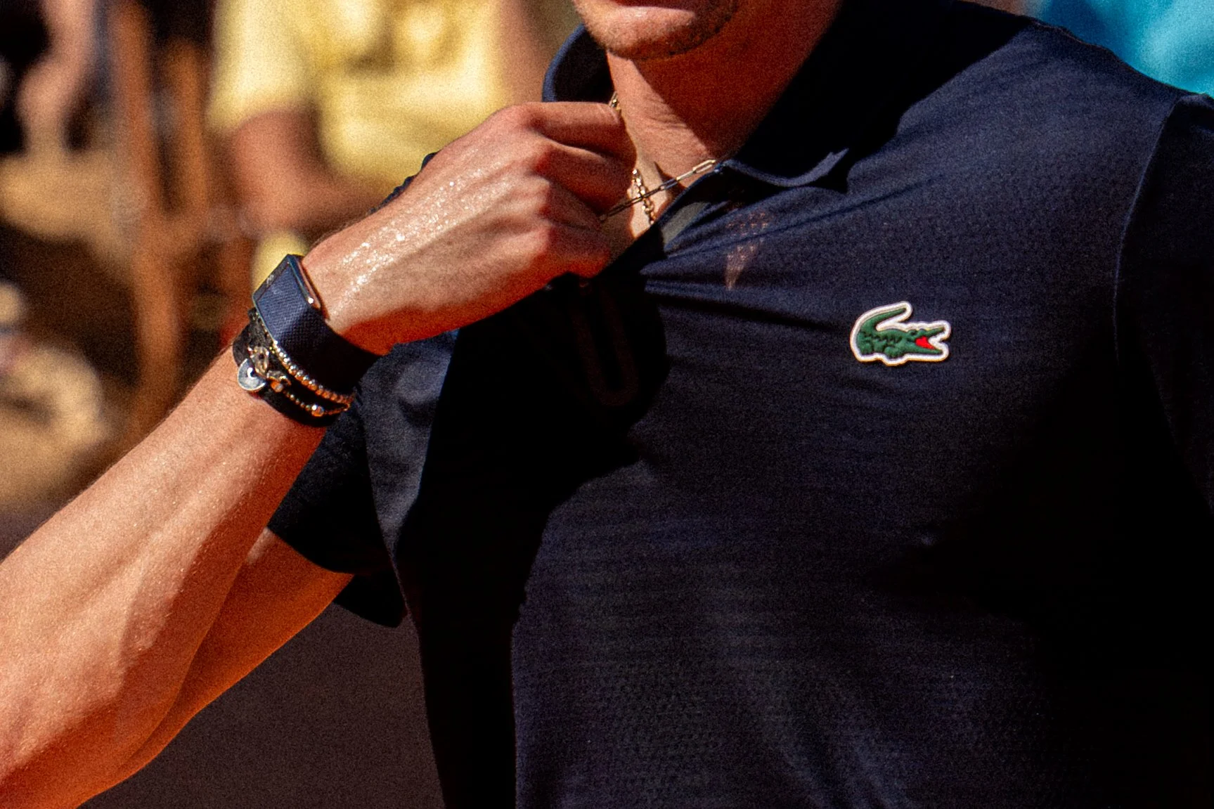 A person wearing a dark Lacoste polo shirt with a crocodile logo, holding a necklace. The person has a bracelet on their wrist and is touching the necklace with their hand.
