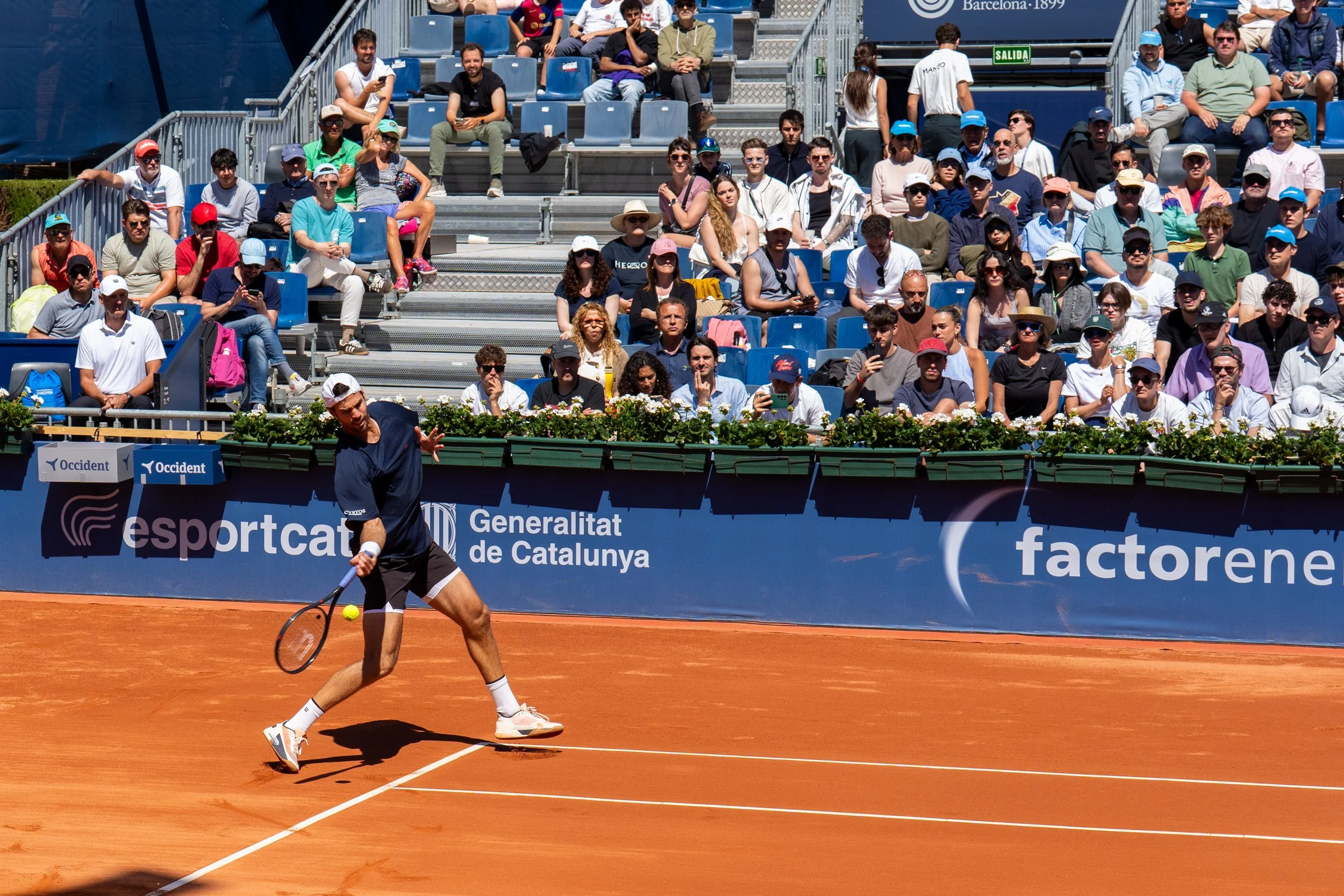 Karen Khachanov on a clay court swinging a racket to hit a yellow tennis ball during a match. Spectators sit in the stands watching, some using their phones. The scene is outdoors on a sunny day.