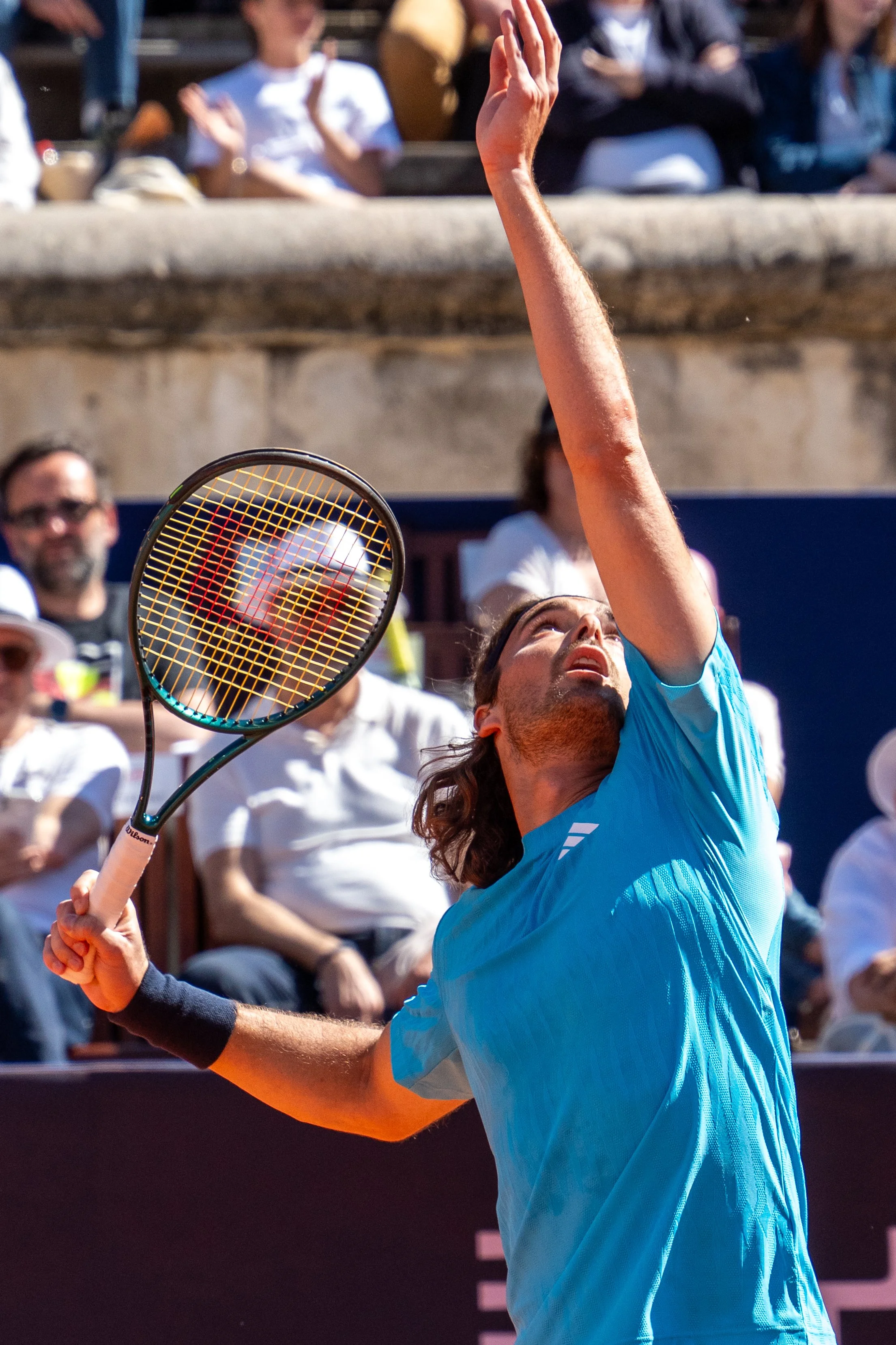 A male tennis player wearing a blue shirt and a black wristband is serving the ball during a match on an outdoor clay court, with spectators sitting in the background.