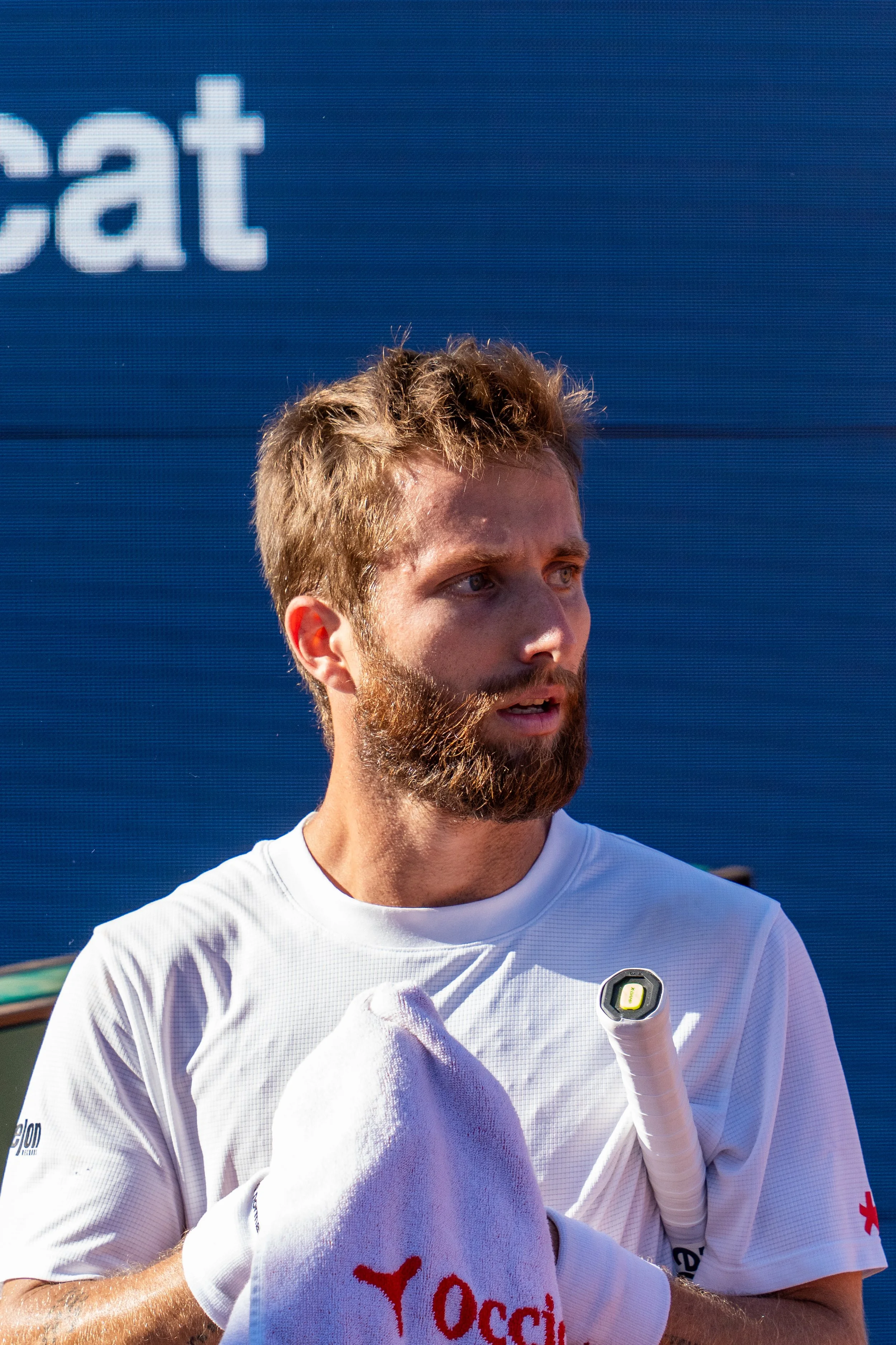 Corentin Moutet holding a white towel with red text and a small flashlight, wearing a white athletic shirt, standing outdoors in front of a blue wall.