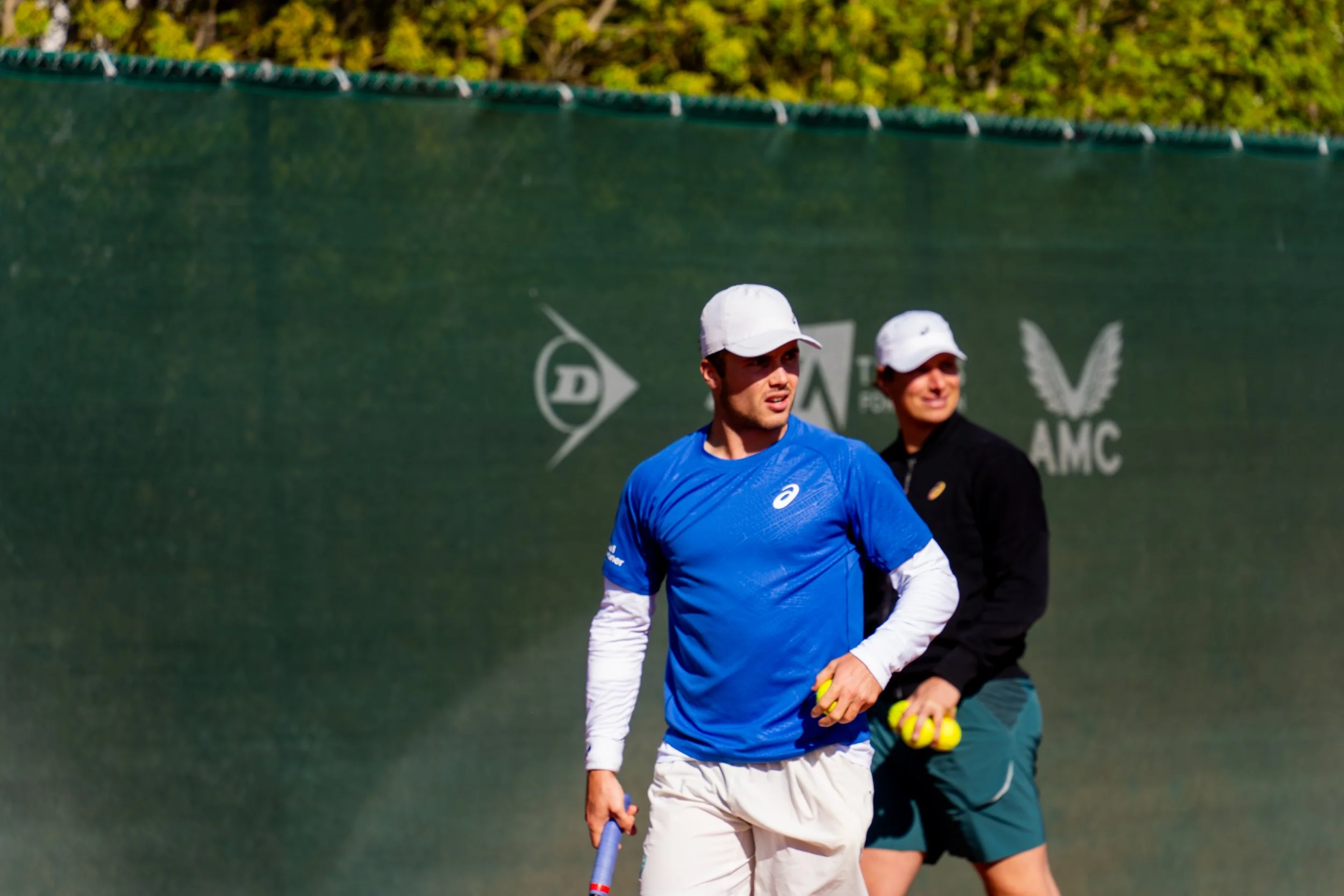 Two people standing on a tennis court holding tennis balls with a green fence and trees in the background.