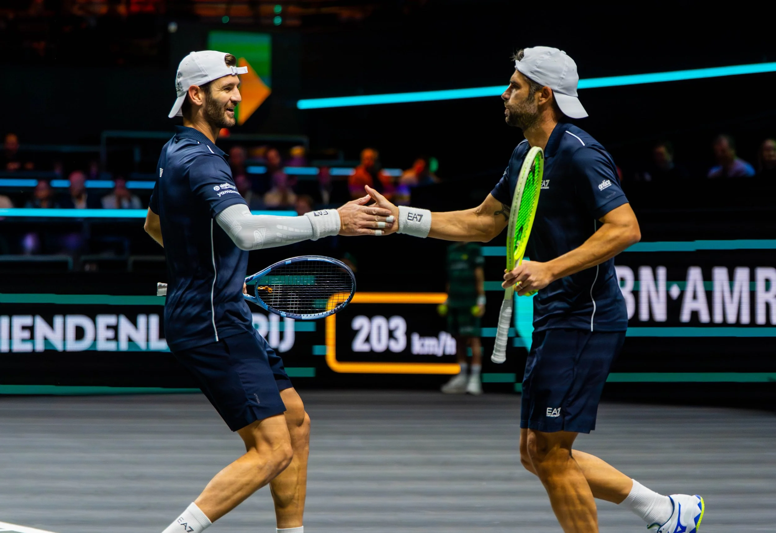 Two male tennis players on a court, dressed in blue uniforms and white caps, shake hands at the end of a match.