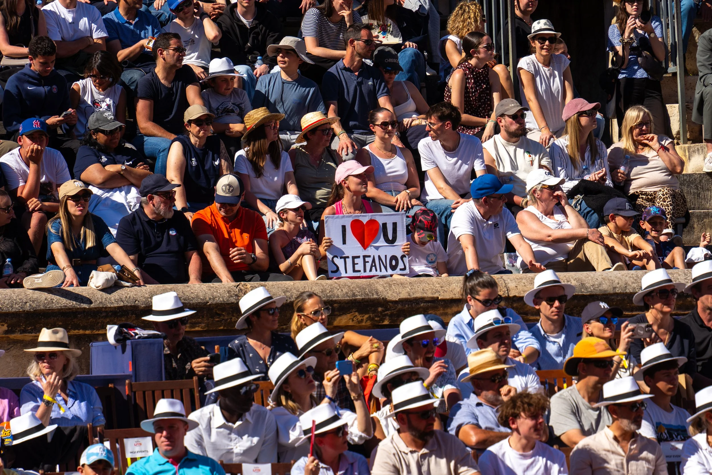 Crowd of people sitting in an outdoor arena, with some holding a sign that says "I ♥ U STEFANOS." Many are wearing hats and sunglasses, enjoying a sunny day.