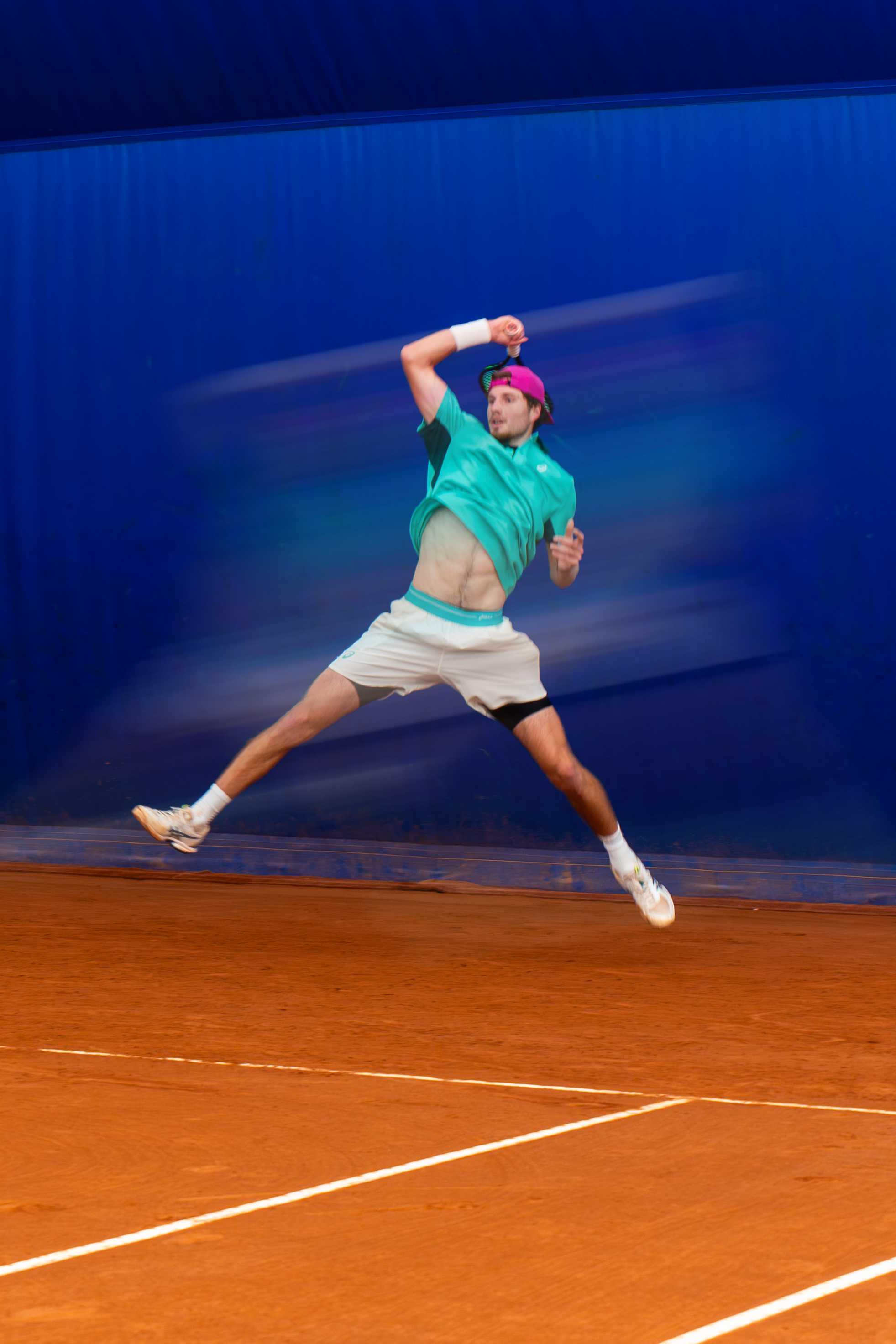 A man playing tennis on a clay court, captured in mid-air as he hits a shot, wearing a purple cap, teal shirt, beige shorts, and white sneakers with black accents.