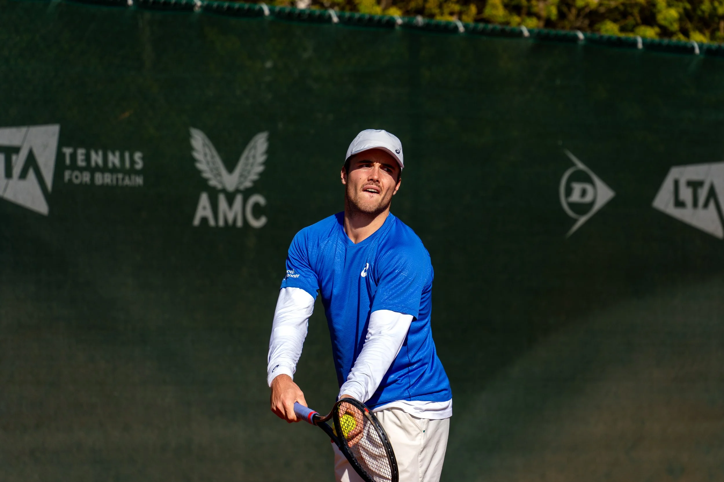 A male tennis player in a blue shirt and white cap playing tennis on an outdoor court.