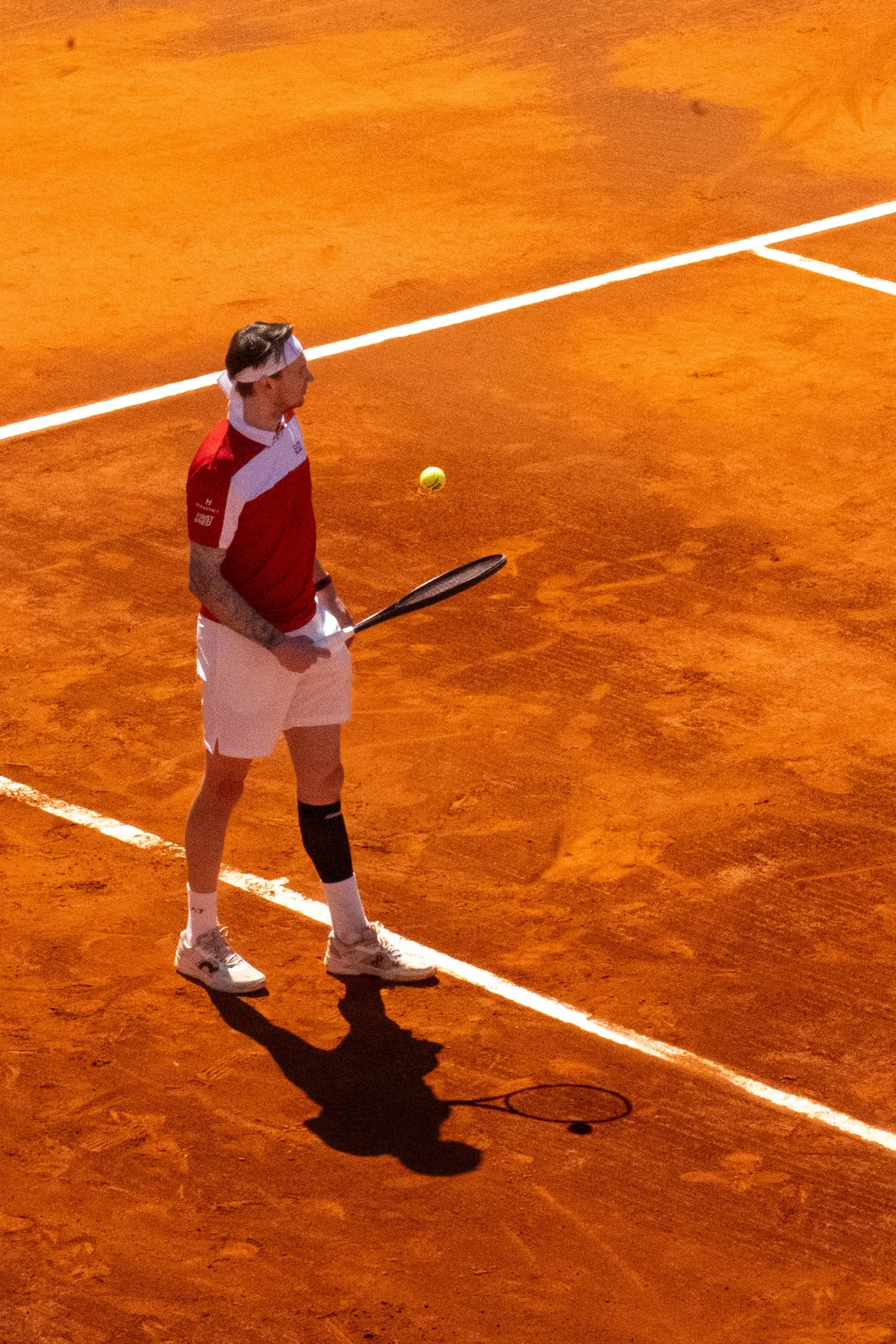 A tennis player on a clay court preparing to hit a tennis ball. The player is wearing a red and white shirt, white shorts, and white tennis shoes, and is holding a tennis racket.