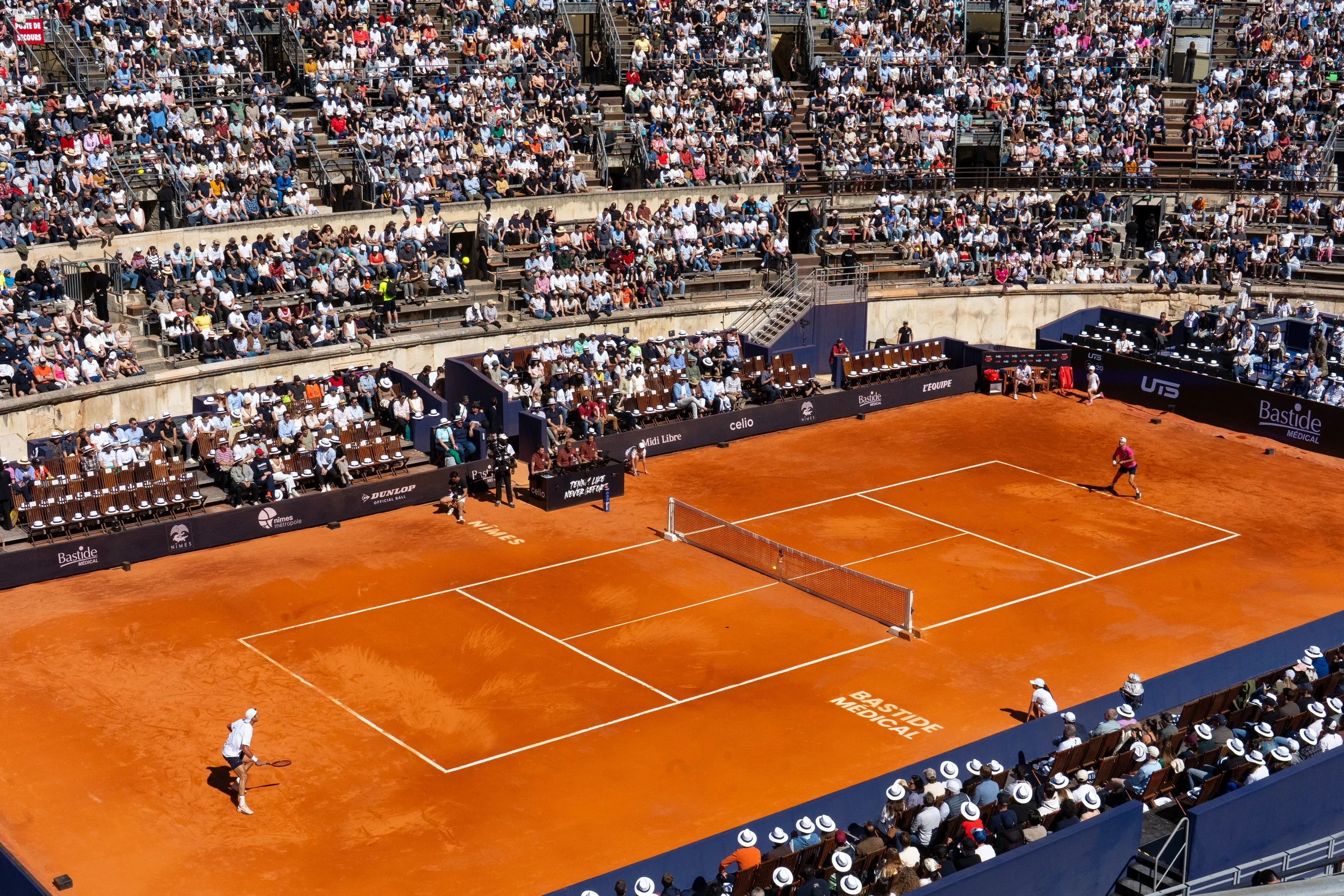 A tennis match on a red clay court with players in action, surrounded by a large audience in the stands.