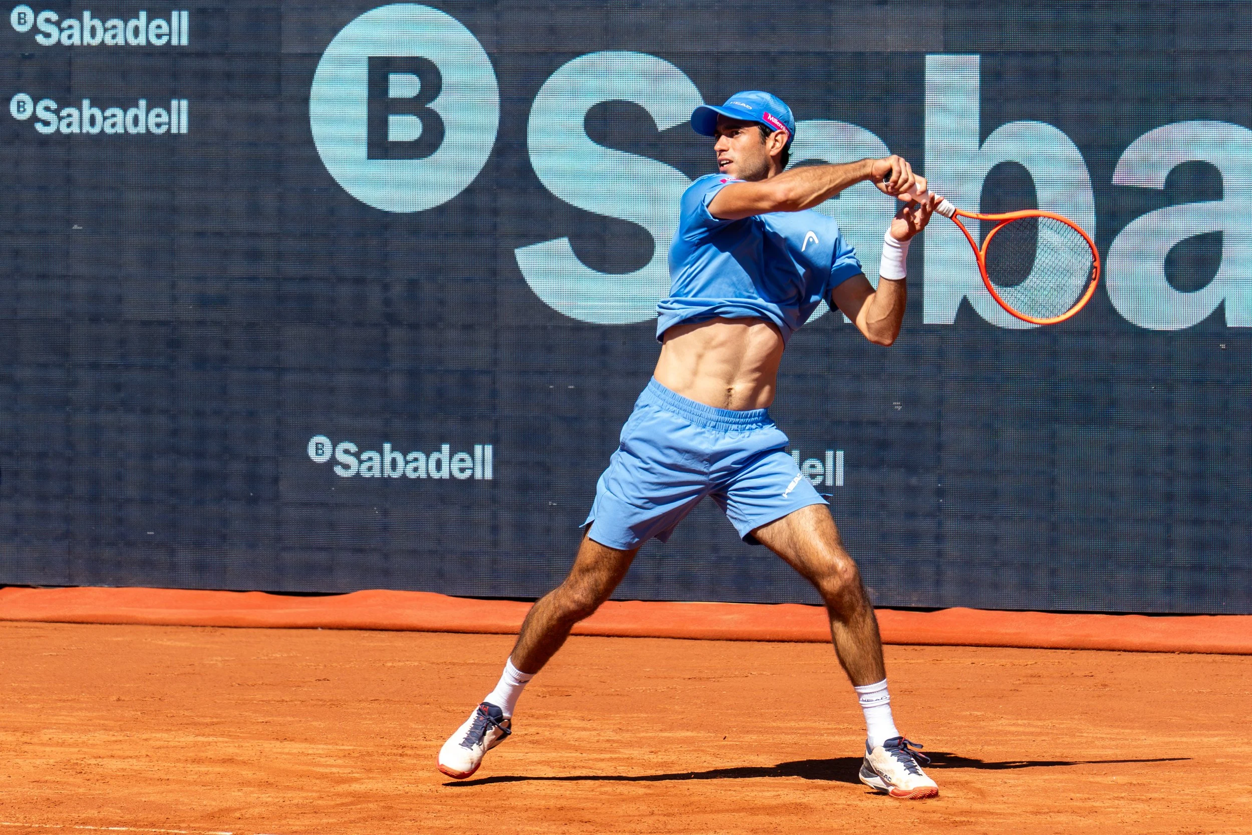 Nuno Borges is preparing to hit a forehand shot on a clay court. He is wearing a blue outfit with a matching cap and shoes, and is holding an orange tennis racket. There are large advertising signs in the background with the logos of Sabadell and B