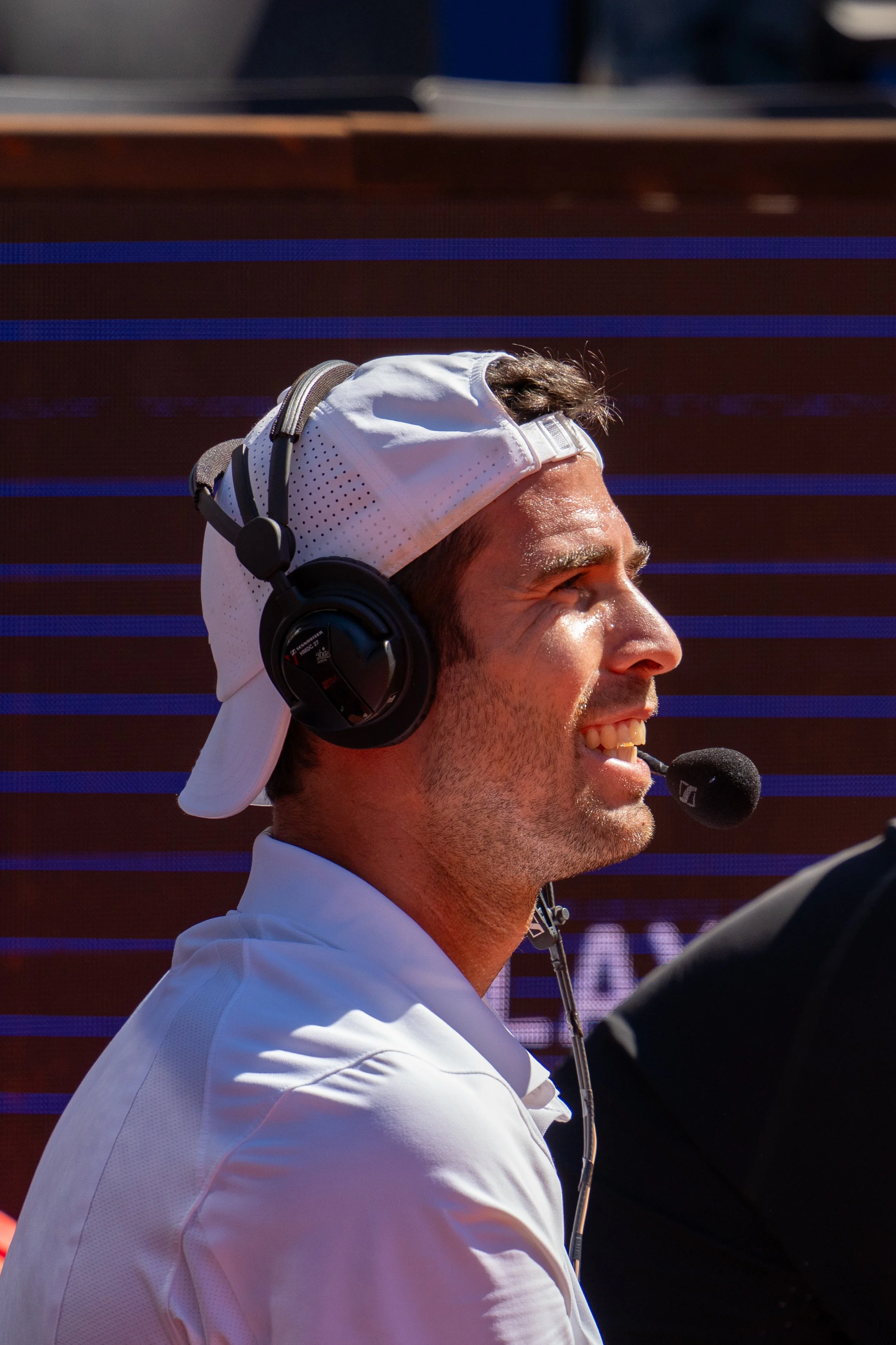 A man wearing a white cap backwards, black headphones, and a white shirt is smiling and talking into a headset microphone during a sports event.