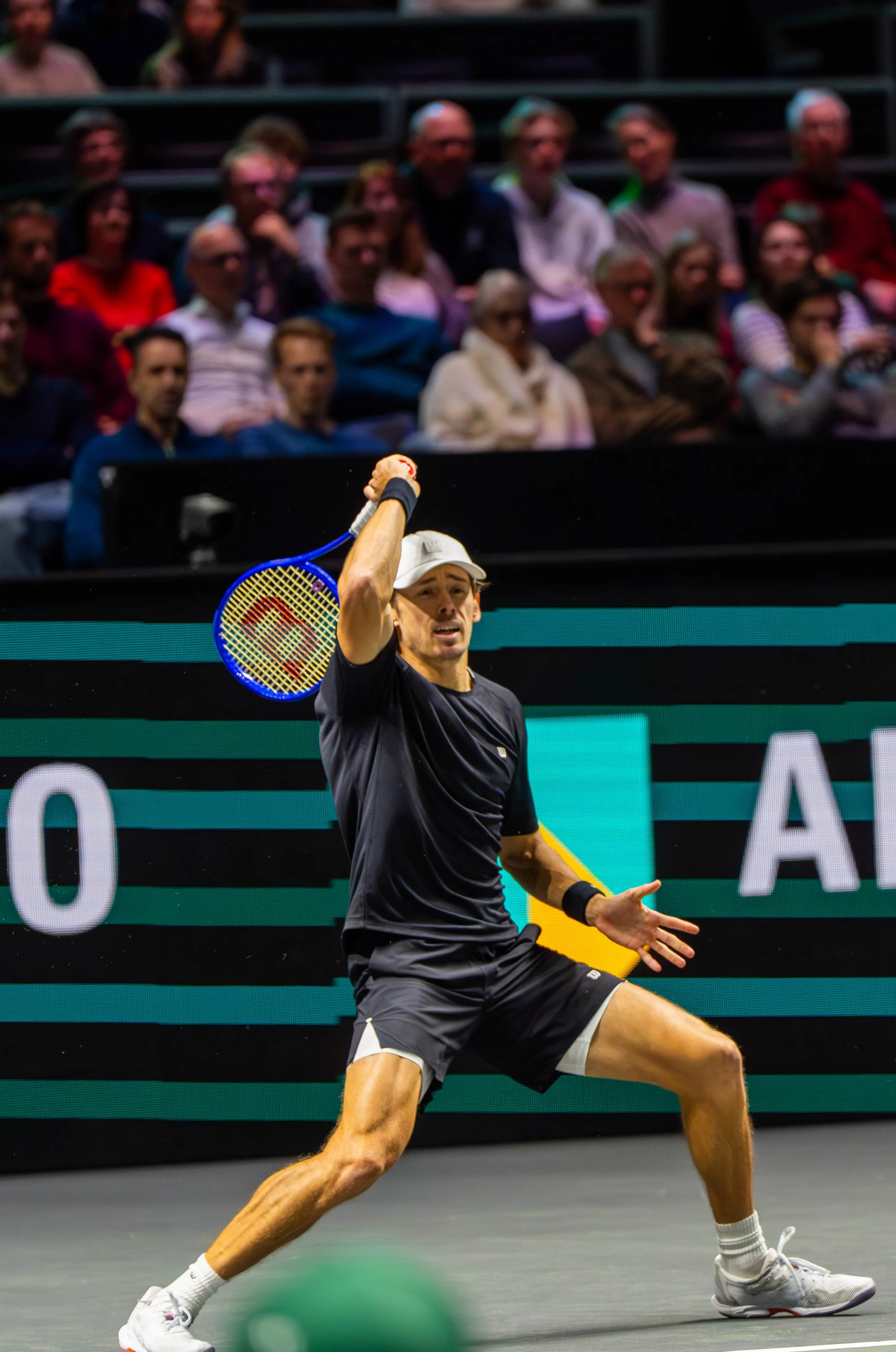 A male tennis player in black attire, white cap, and white shoes practicing a tennis shot during a match on an indoor court, with spectators watching intently in the background.